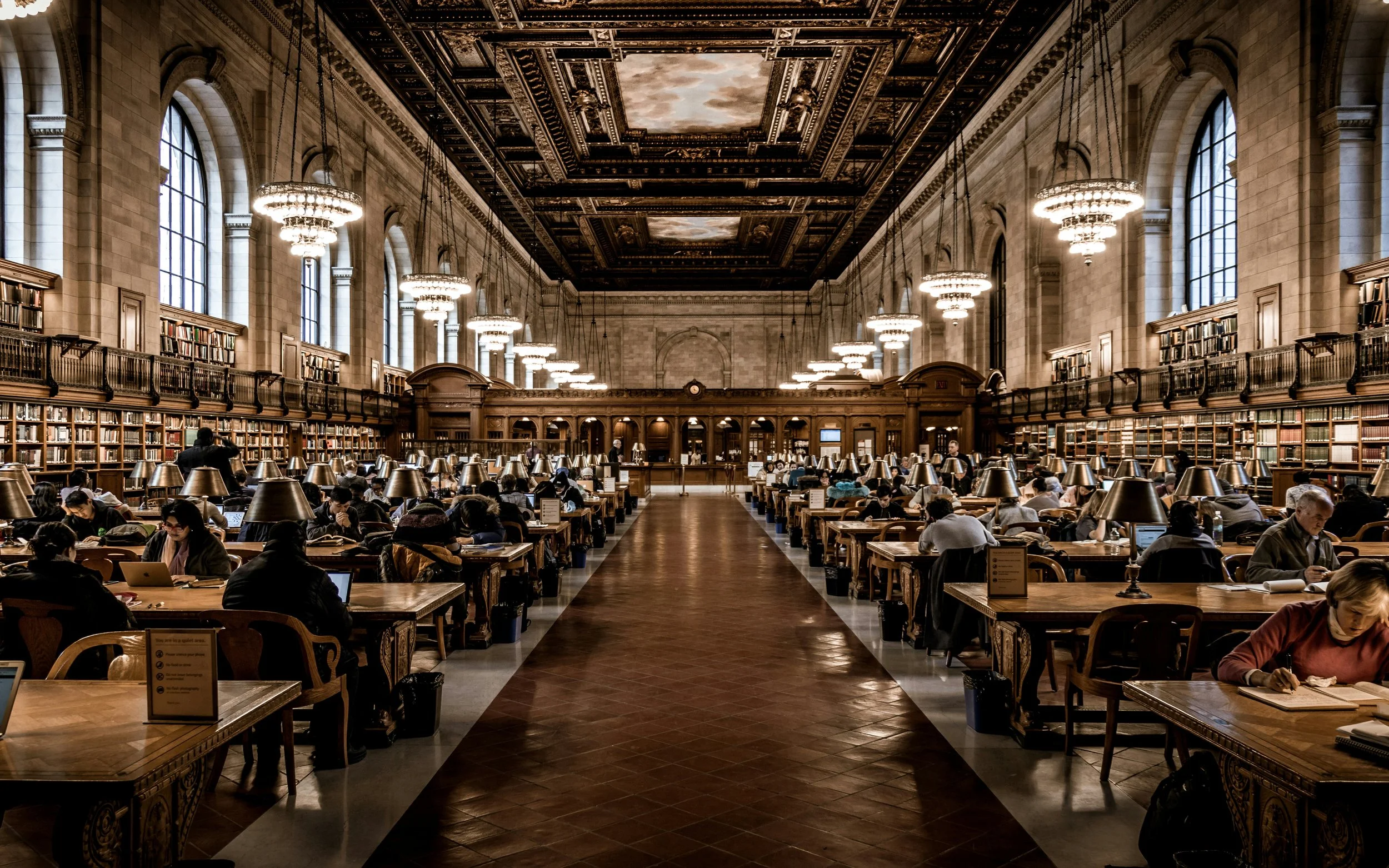 Inside a grand historic library with tall arched windows, multiple chandeliers hanging from a decorated ceiling, and high bookshelves, with patrons reading and working at wooden tables equipped with individual lamps.