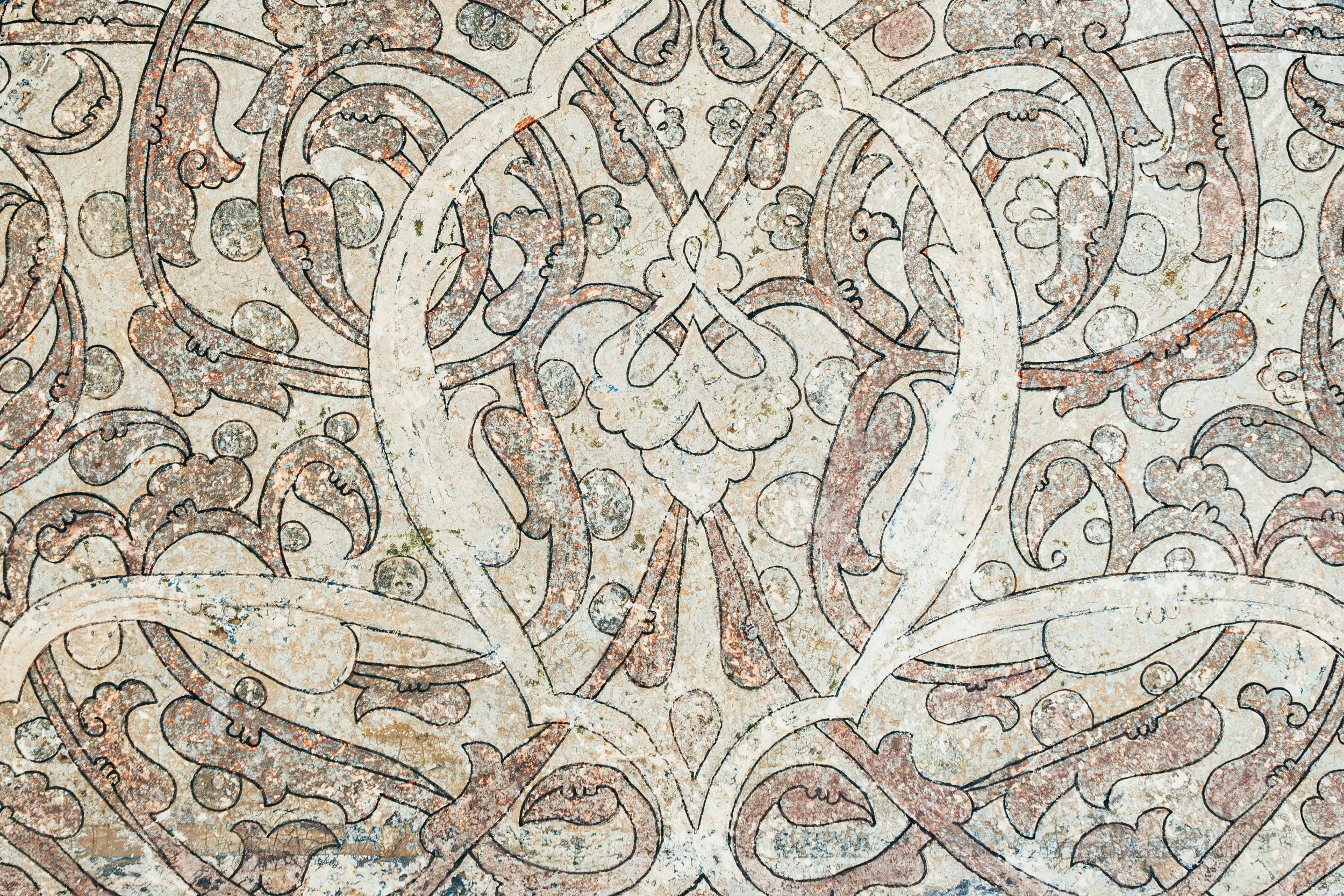 Close-up of an intricate, weathered wall with detailed geometric and floral patterns in faded beige, brown, and reddish tones.