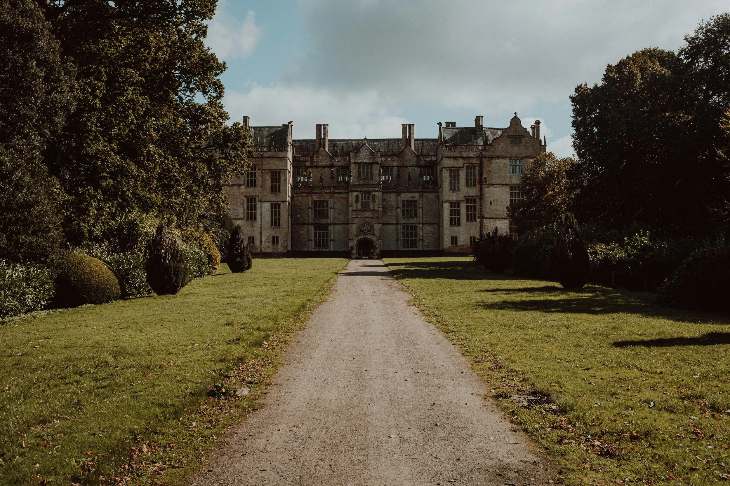 A long straight gravel pathway leading towards a large historic castle or mansion surrounded by well-maintained lawns and tall trees on a cloudy day.