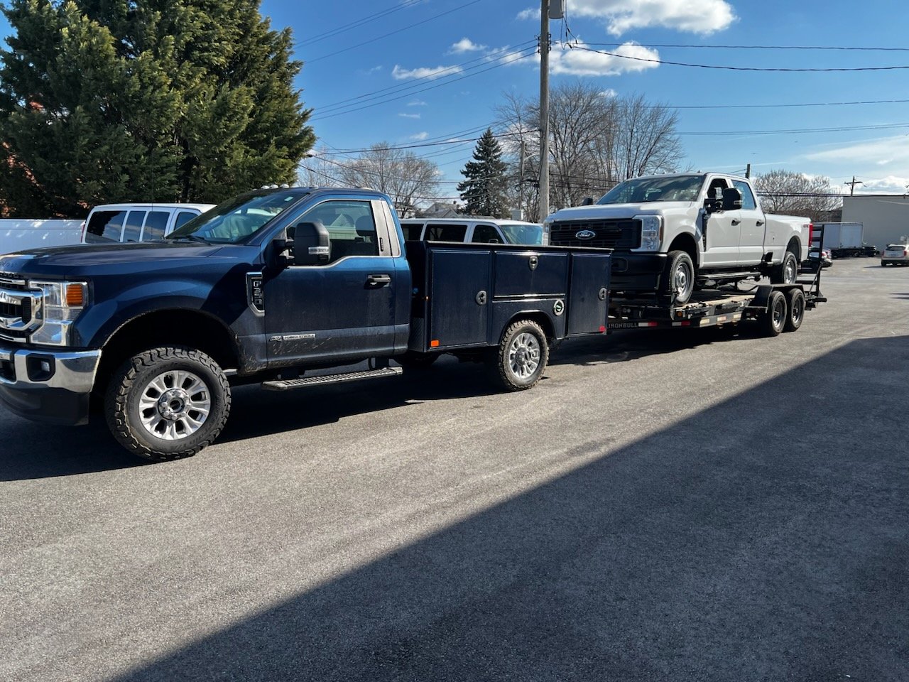 Black pickup truck towing a white pickup truck on a flatbed trailer in an outdoor parking lot on a clear day.