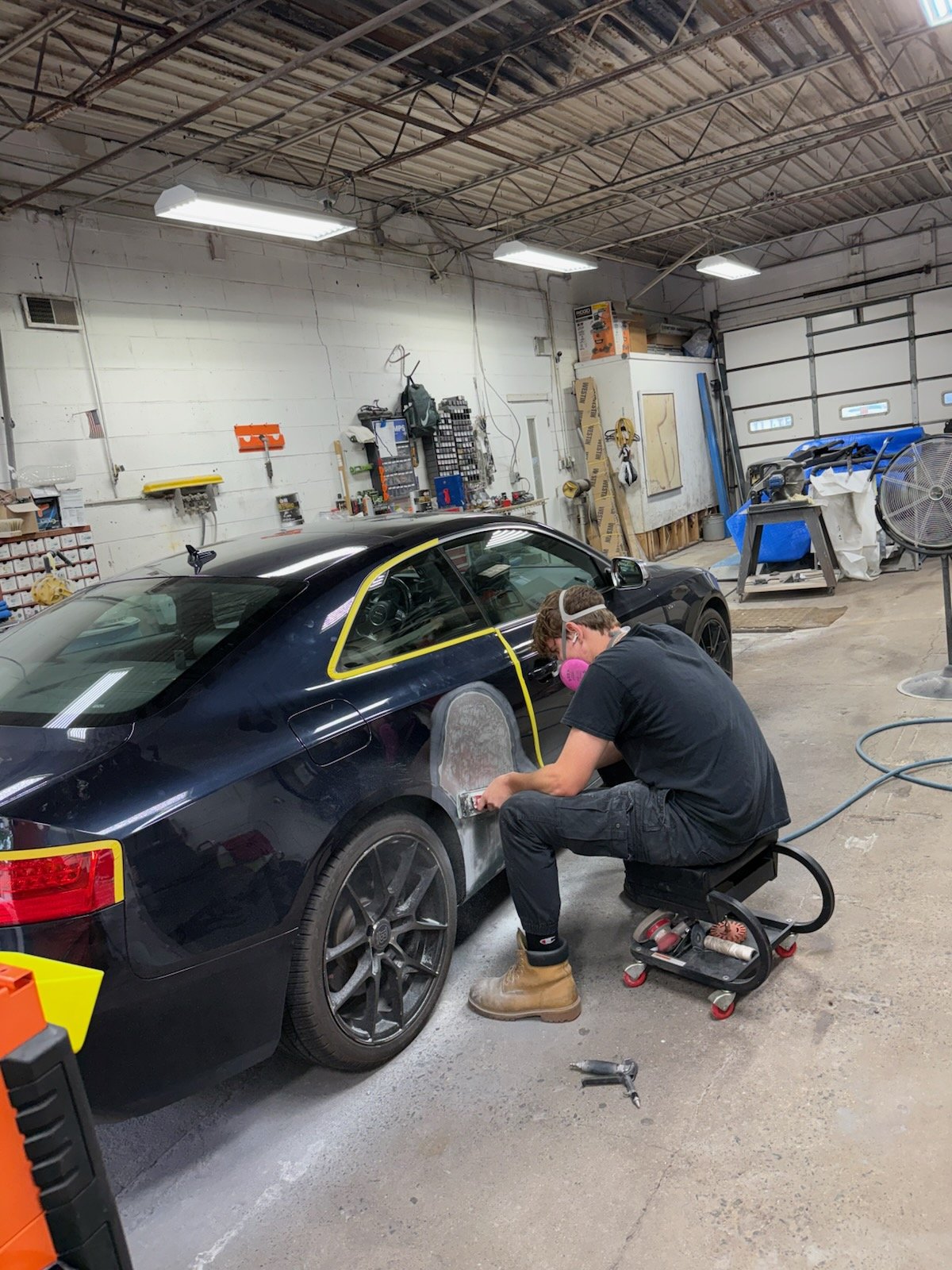 A person in safety gear polishing a black sports car inside an automotive workshop or garage. The workshop has tools, equipment, and materials on shelves and walls, with a large garage door in the background.