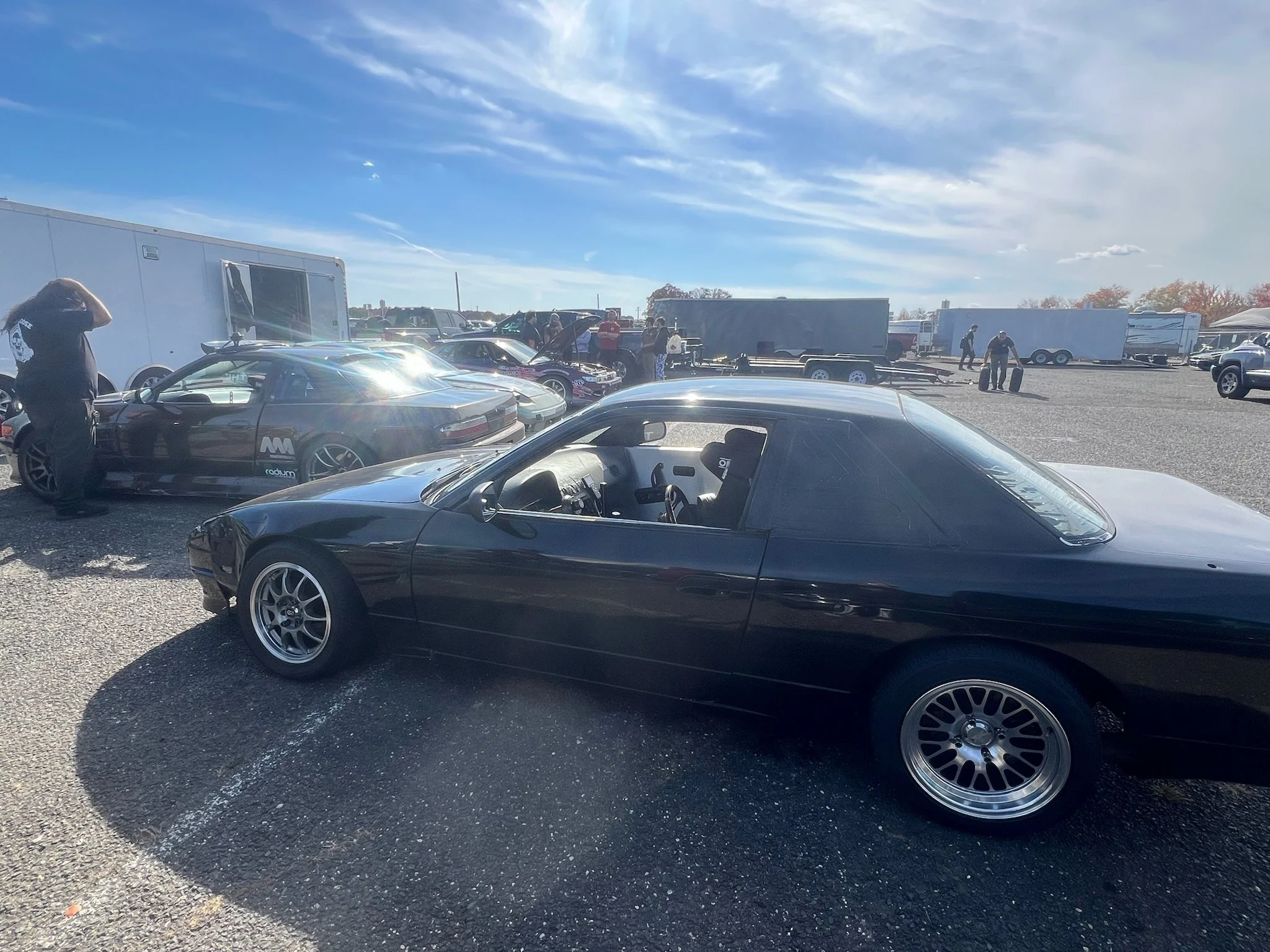 View of a parking lot with multiple cars, several people around, and trailers, under a partly cloudy sky.