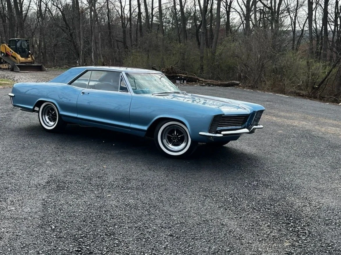 A vintage blue classic car parked on gravel ground with a wooded background and construction equipment in the distance.