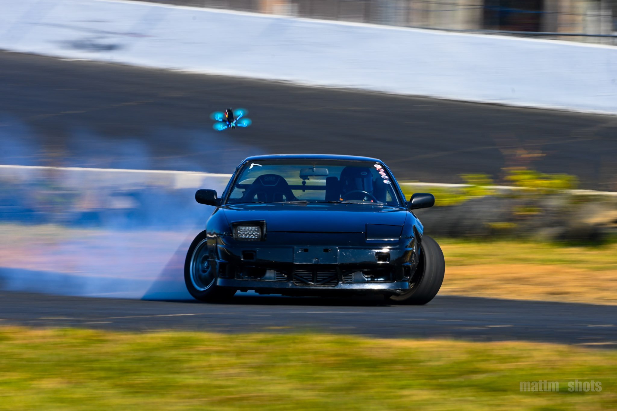 A black sports car drifting on a race track, with a drone flying nearby capturing the action. The car is kicking up dust and appears to be in a controlled slide.