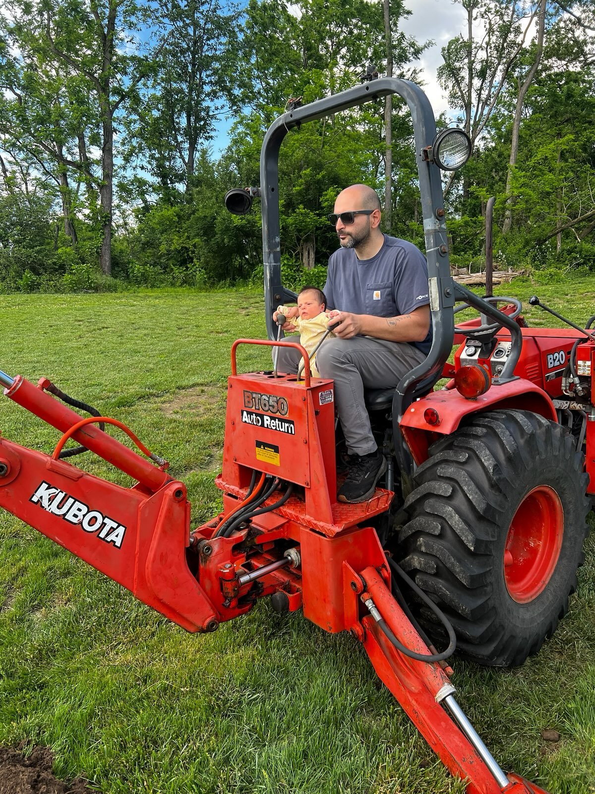 A man with a beard wearing sunglasses and a gray t-shirt sitting on a red Kubota tractor, holding a small baby or toddler in a yellow outfit, outdoors on a grassy field with trees in the background.
