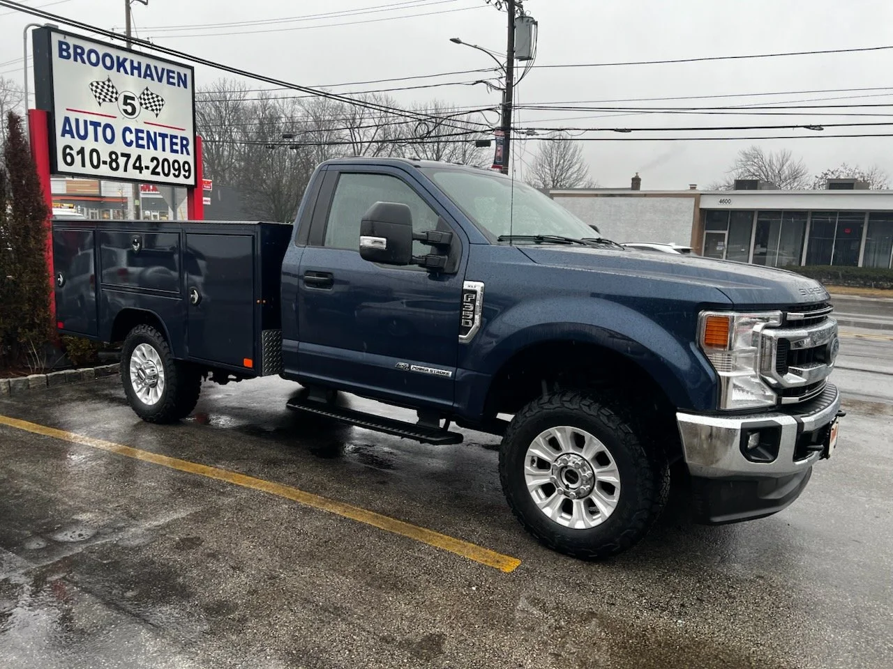A blue Ford F-250 pickup truck parked in a wet parking lot near a sign for Brookhaven Auto Center, with a commercial building in the background and power lines overhead.