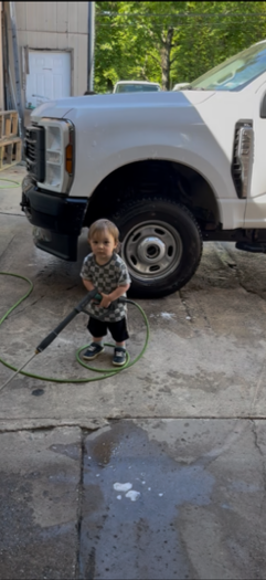 A young boy playing with a garden hose near a large white truck parked outside a building with a metal wall in a lush green area.