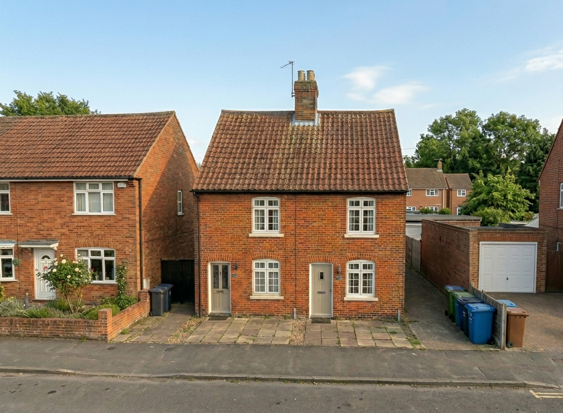 Two-story brick house with a red tiled roof, white window frames, and a front door on a paved driveway, next to a similar house, with a garage and green bins on the side.