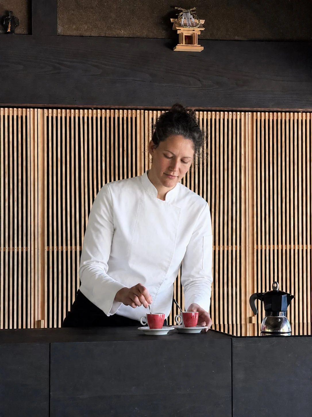 白い制服の女性がコーヒーショップのカウンターでコーヒーを準備している。背景は木のパネル.