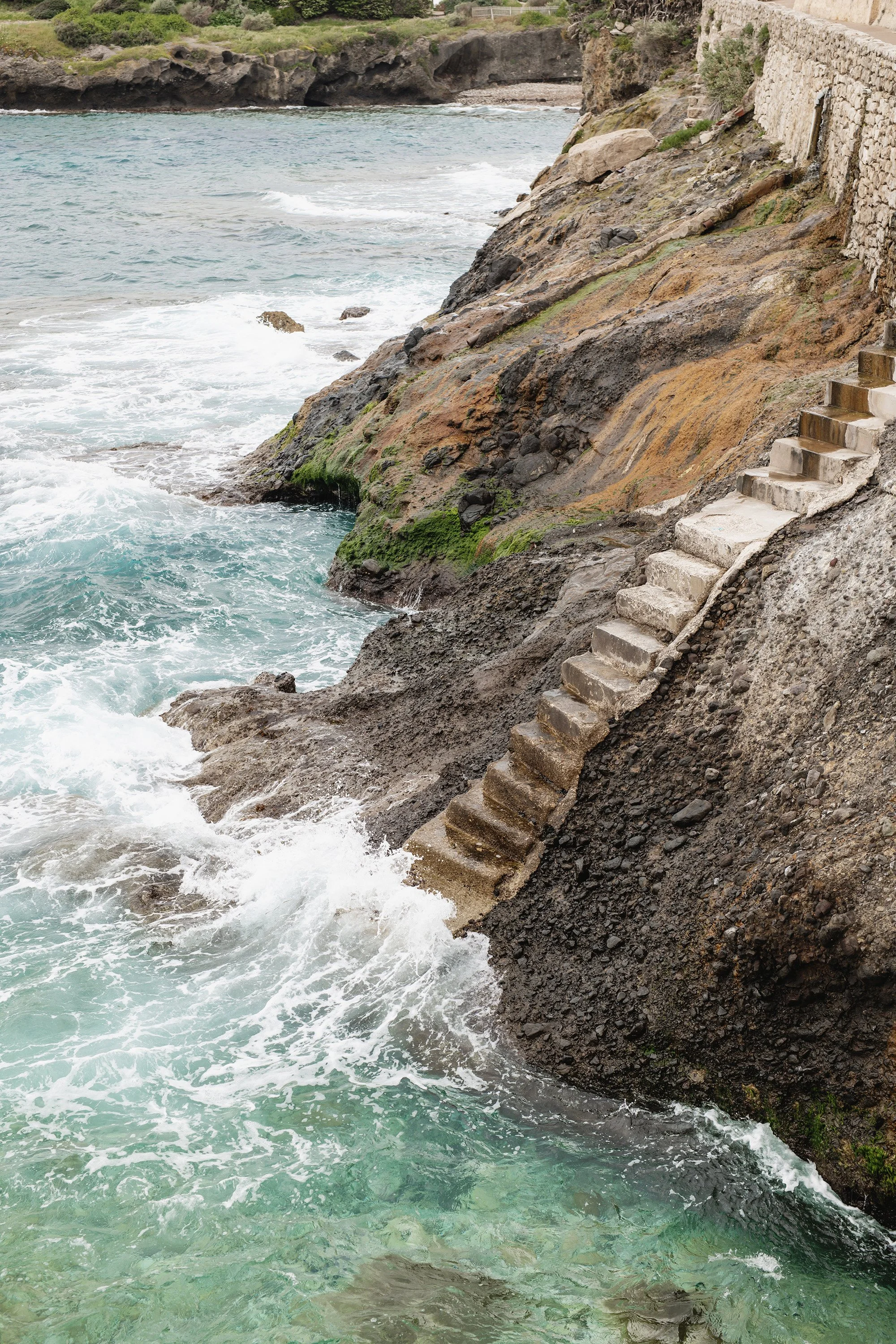 Concrete stairs leading down to a rocky shoreline with ocean waves crashing against the rocks, with cliffs and greenery in the background.