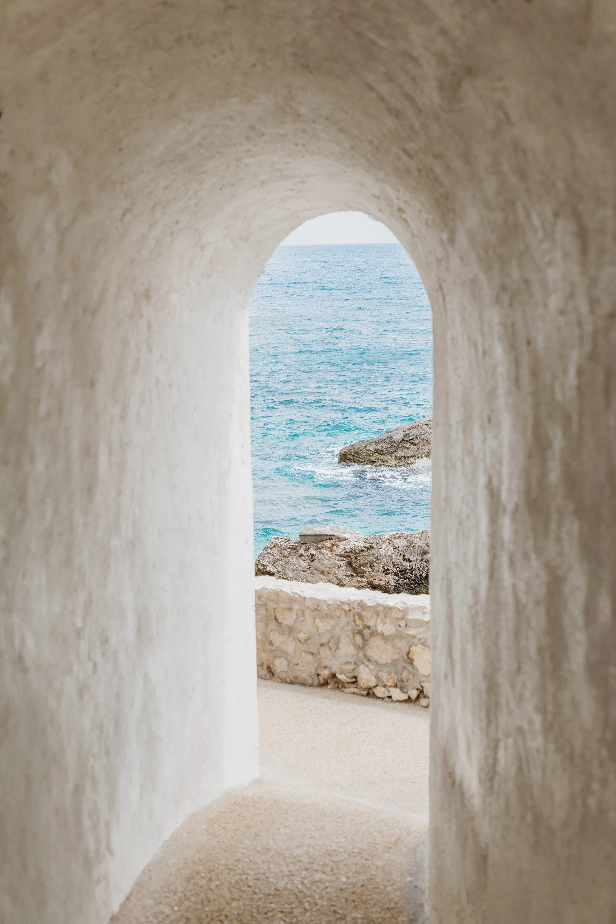 View of the ocean and rocks through an arched opening in a white stucco wall.