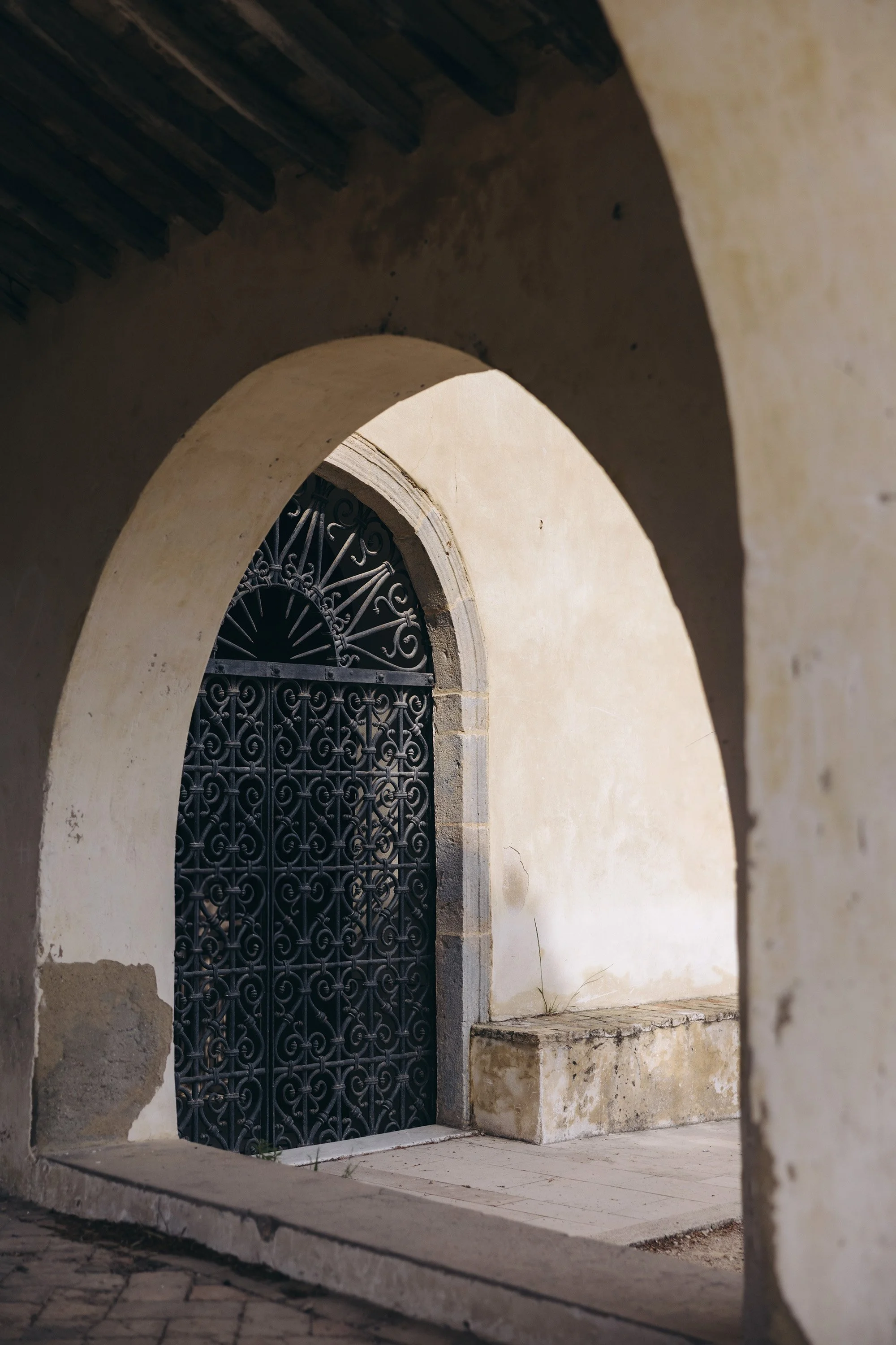An arched stone doorway with a decorative black wrought iron gate, set in an aged beige wall with a stone bench nearby.
