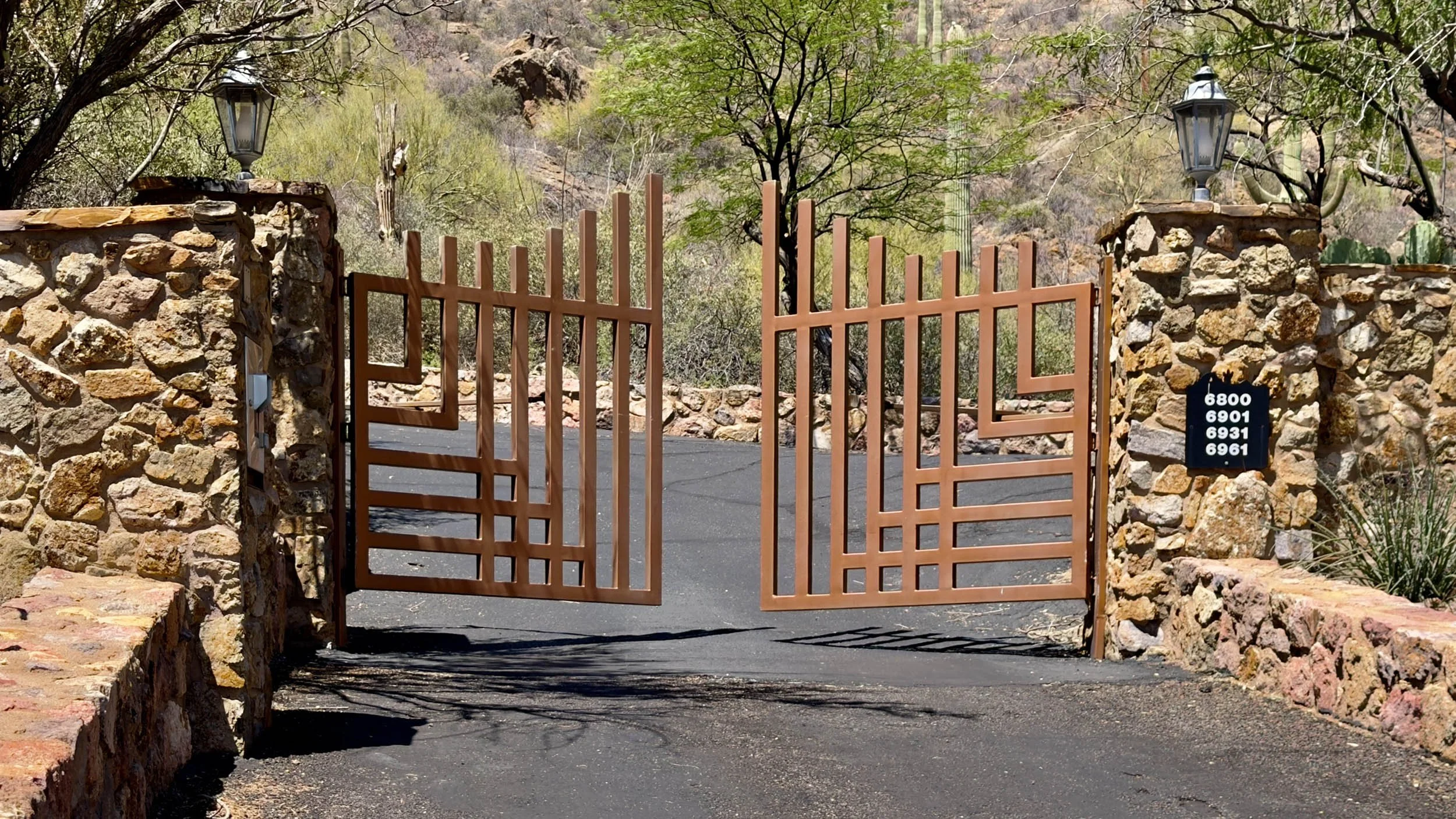 An open metal gate with a geometric pattern at a driveway entrance, flanked by stone walls with lanterns on top, set against a desert landscape with trees and rocky hills in the background.