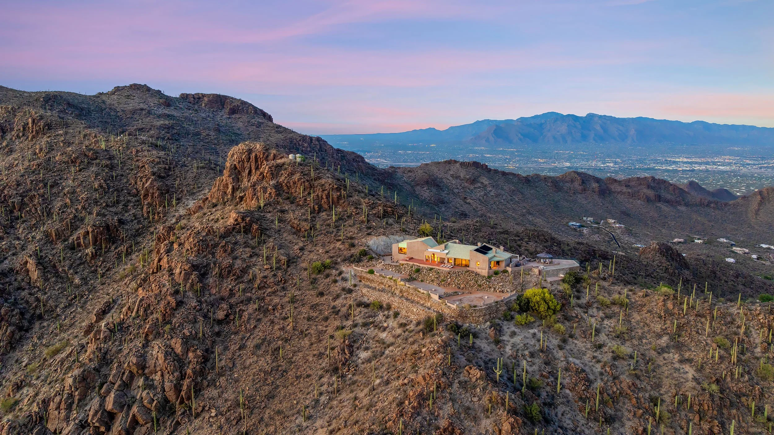 A house situated on a rocky hillside with cacti, overlooking a city and mountain range at sunset.