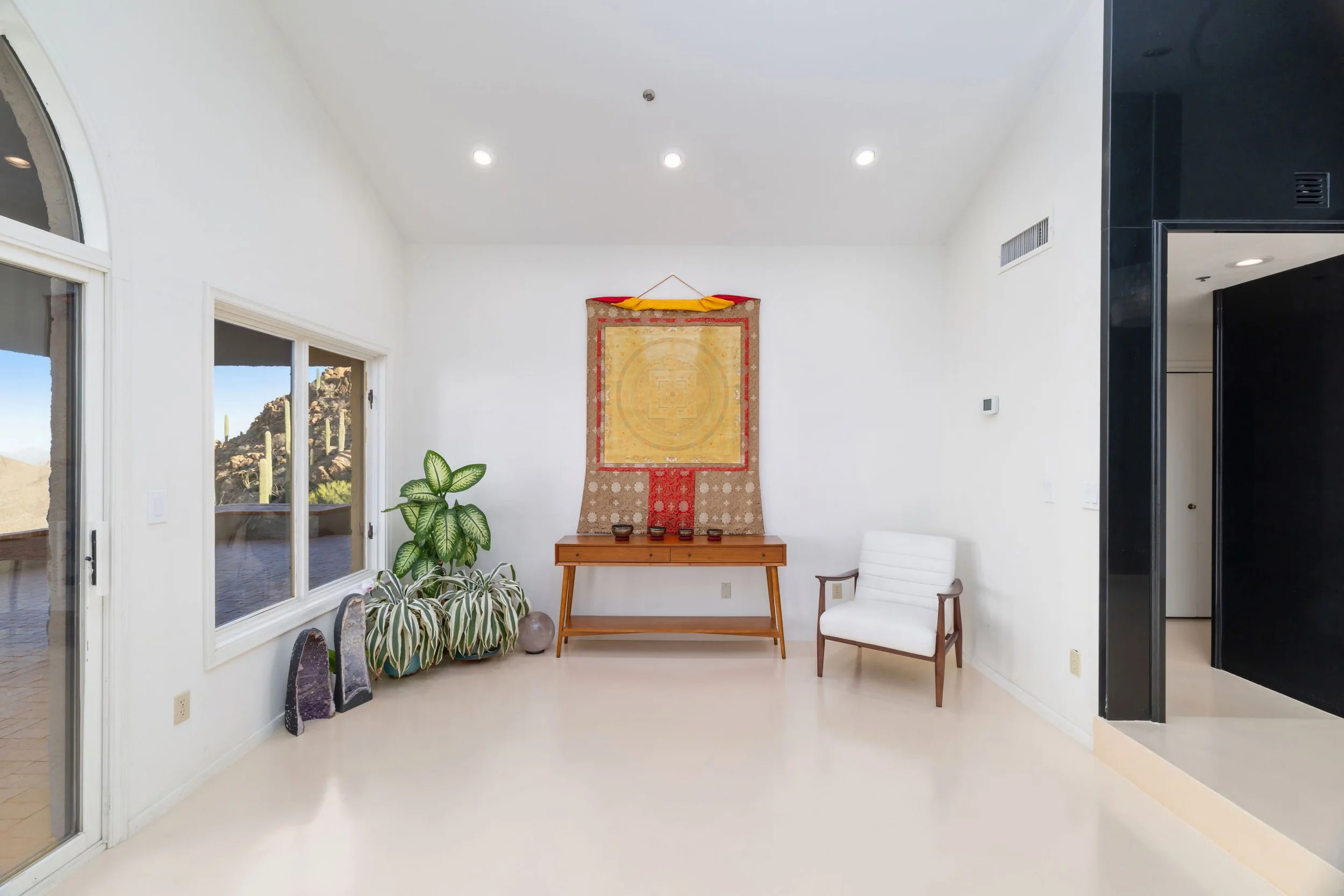 Interior view of a modern room with white walls and ceiling, featuring a wall hanging artwork, a wooden console table with small decorative items, a large houseplant, a white armchair, and windows showing a desert landscape outside.