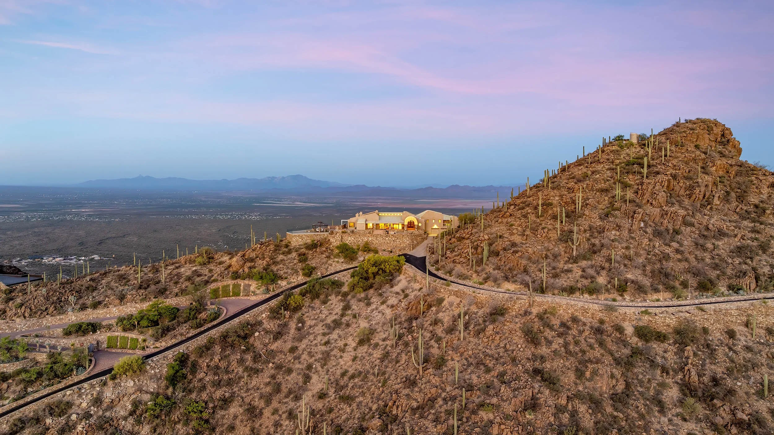 A house built into a rocky hillside with desert plants and cacti, overlooking a vast desert landscape and mountains in the distance at sunset.