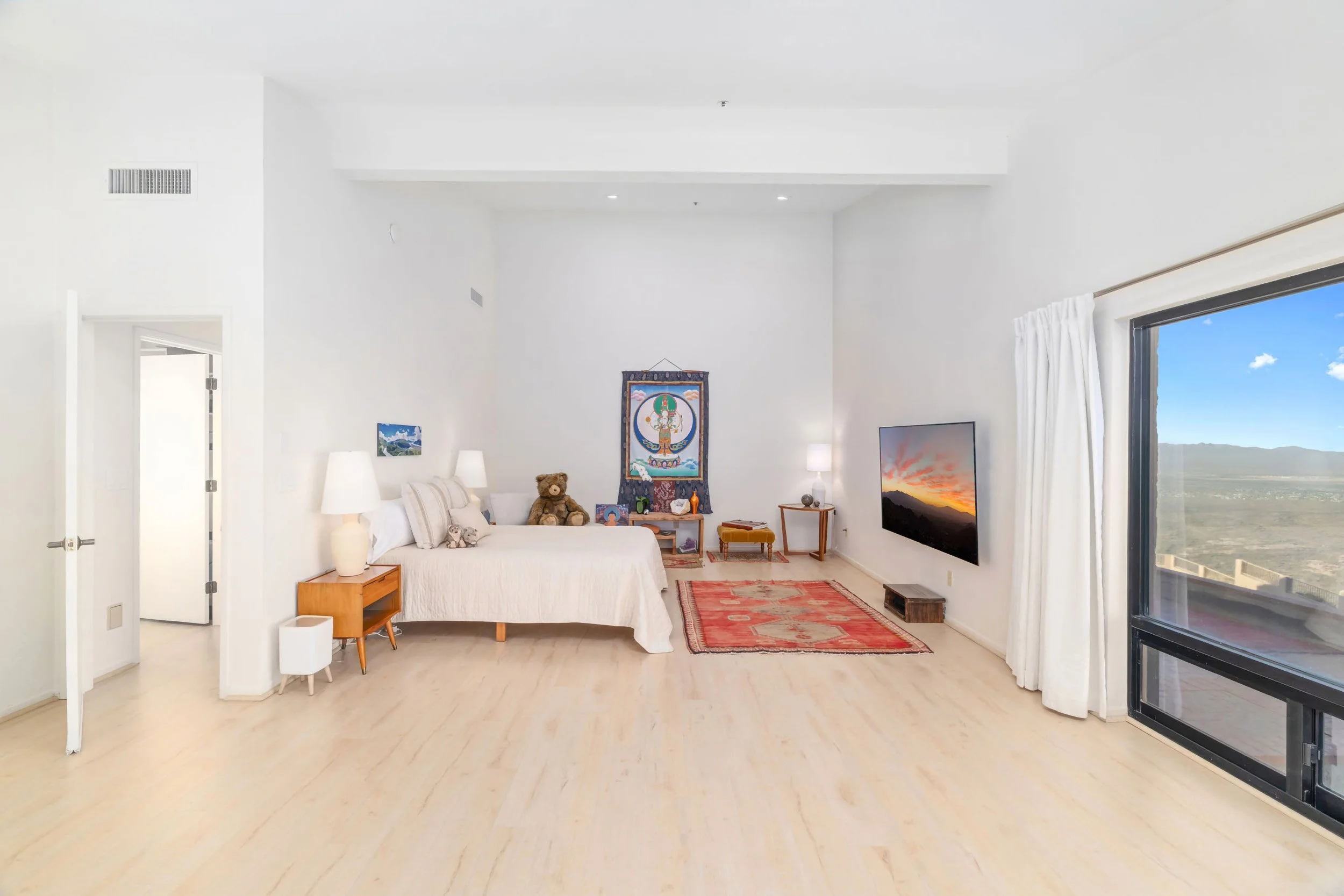 Living room with white walls, large sliding glass door, bed with pillows, teddy bear, Asian artwork, television, and window with a view of mountains and blue sky.