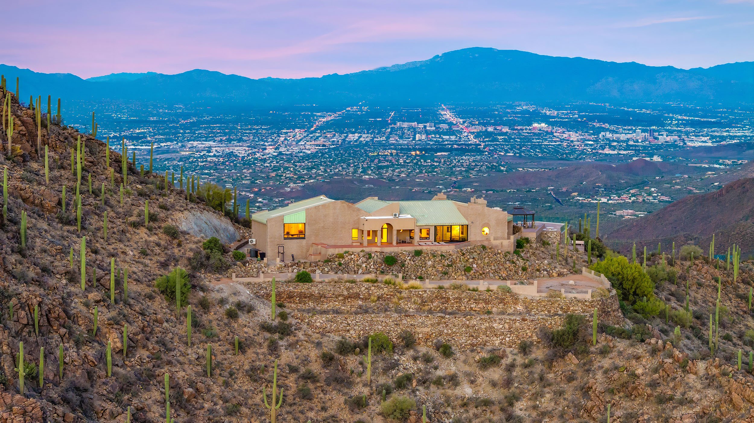 A house on a hillside overlooking a city with mountains in the distance, desert vegetation, and cacti surrounding the house.