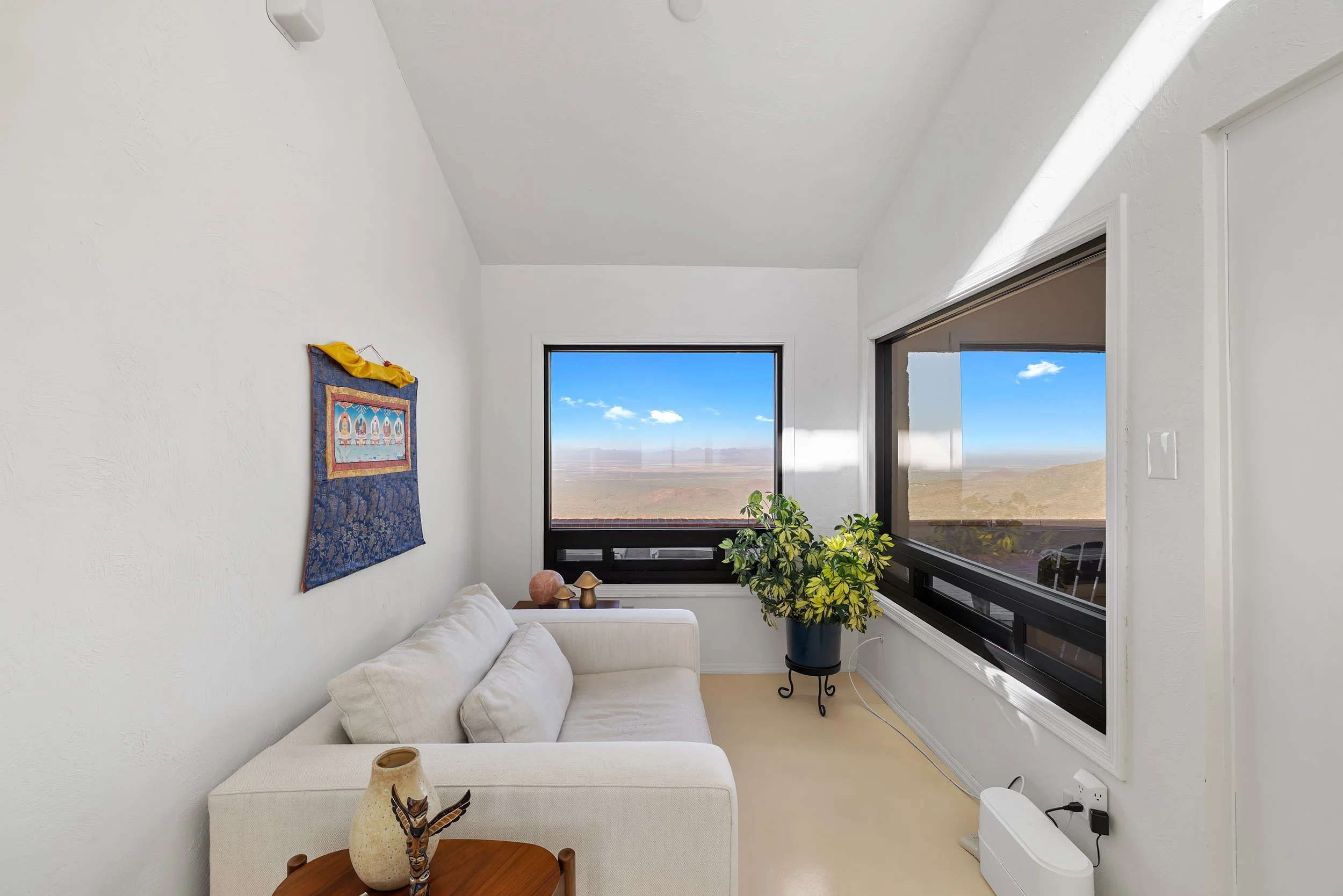 Cozy living room with a white sofa, a wooden side table with a decorative vase, and a large window showing a desert landscape and blue sky. Plants and artwork adorn the room, with natural light illuminating the space.