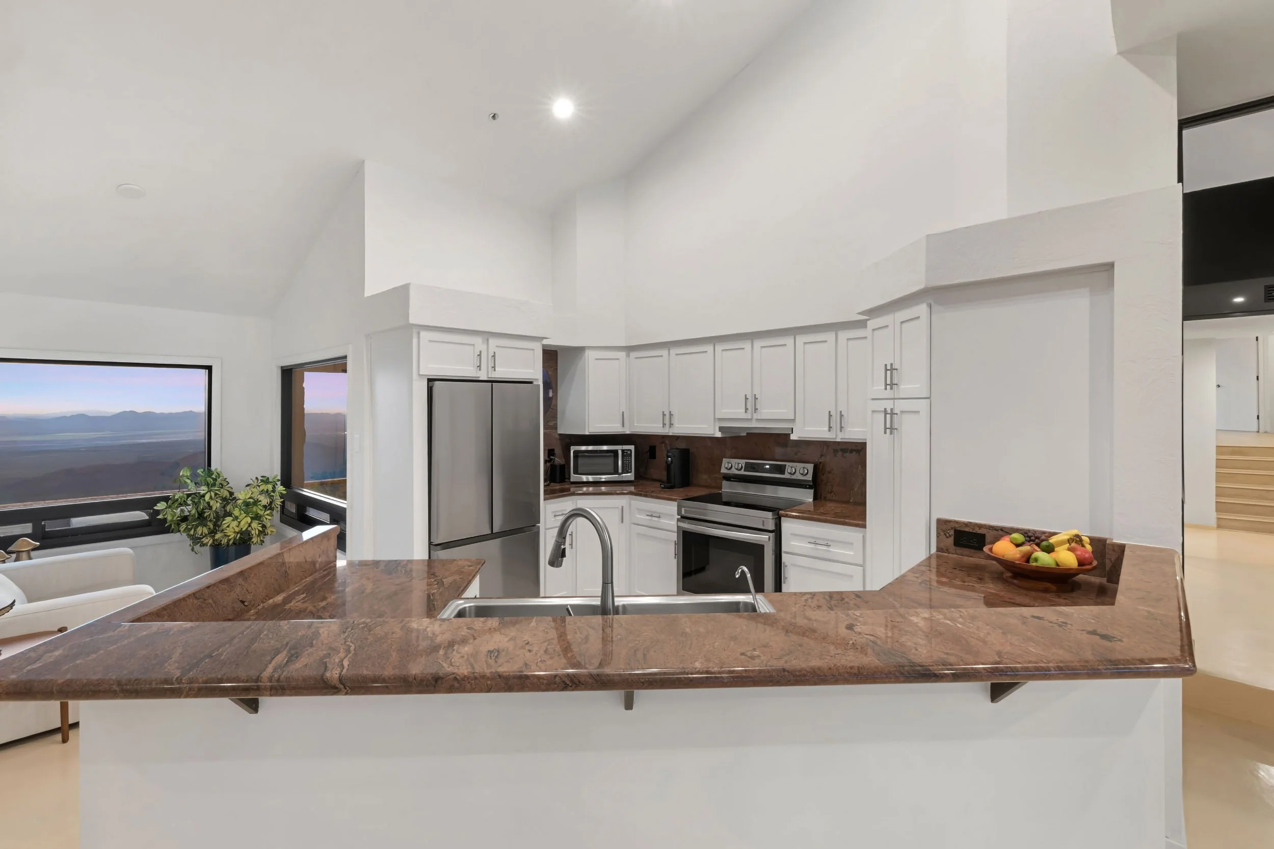 Kitchen with white cabinets, stainless steel refrigerator, microwave, oven, and brown granite countertops. A window shows a mountain view, and there is a bowl of colorful fruit on the counter.
