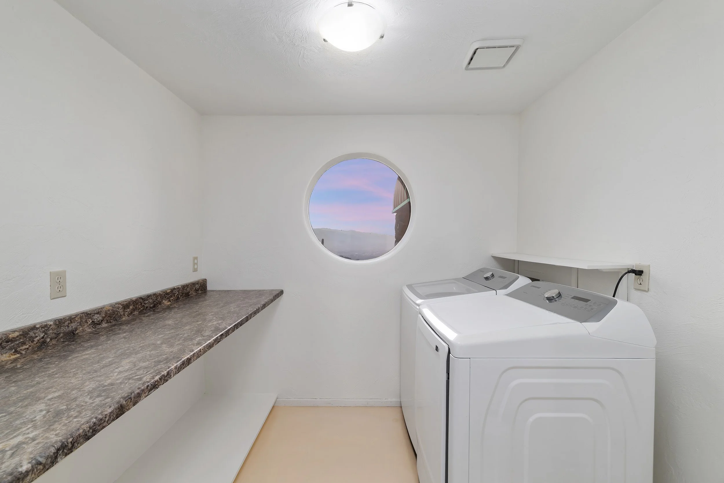 A laundry room with white walls, a round window showing a sunset sky, a brown granite countertop, and a white top-loading washing machine.