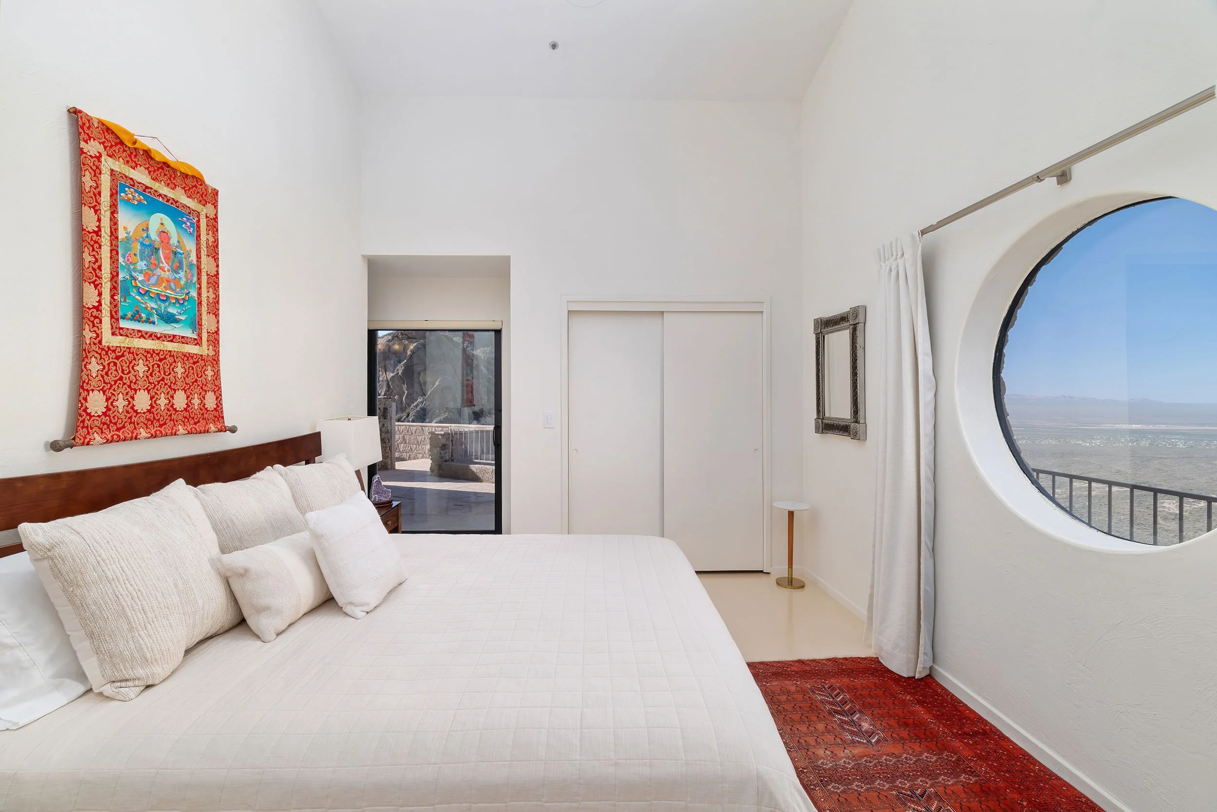 A bedroom with a white bed and pillows, a red patterned rug, a round window showing an outdoor landscape, and a mountain view.