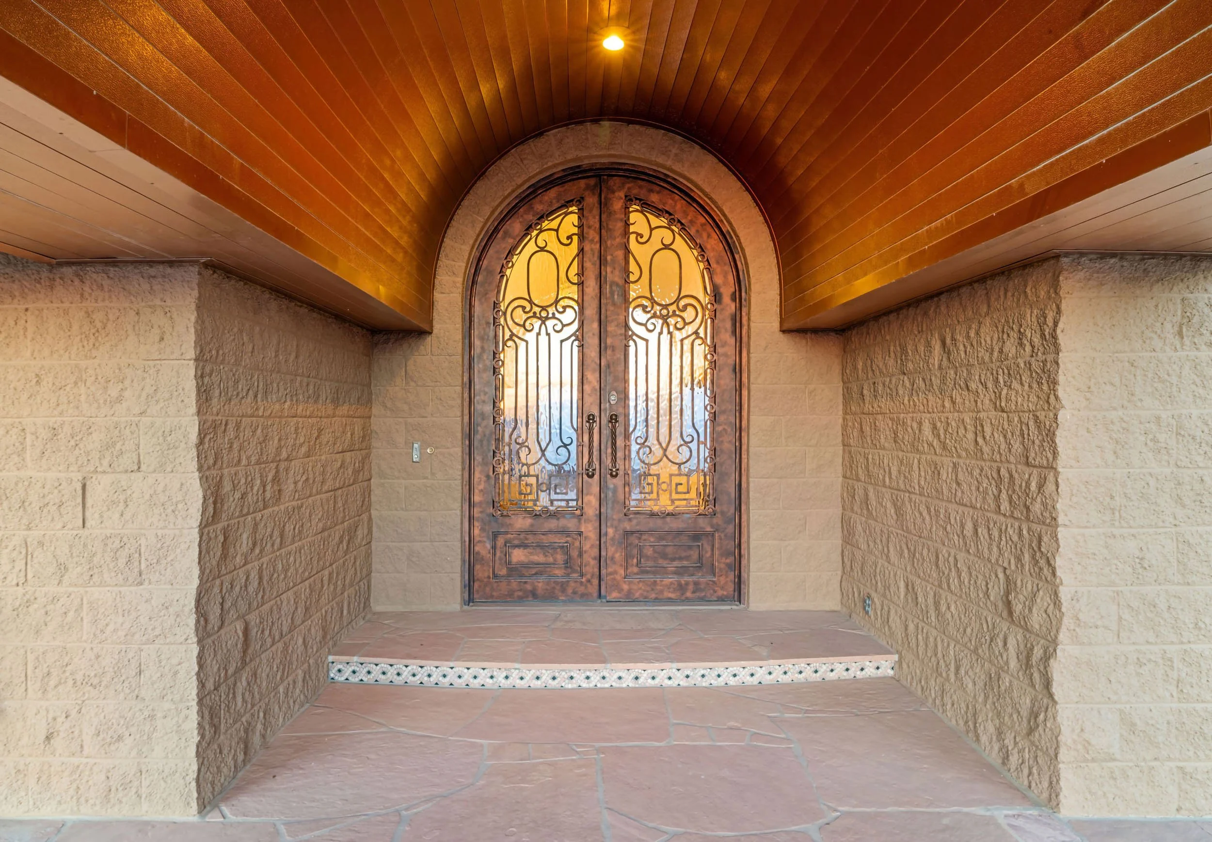 Front door with ornate ironwork, surrounded by stone walls and a wooden overhang at a porch entrance.