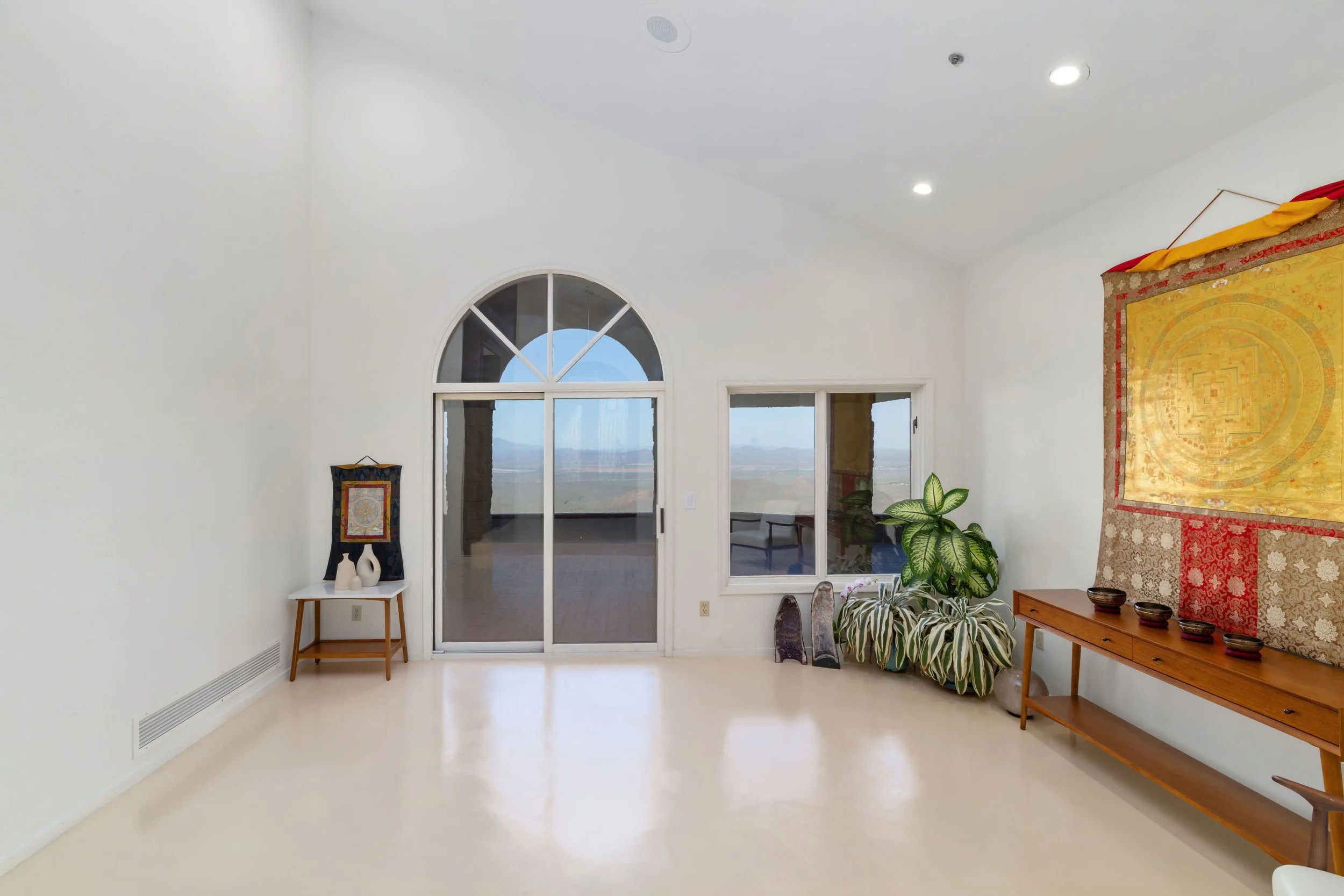 A spacious living room with a high ceiling, white walls, and large arched windows providing a view outside. Decor includes a small white table with vases, a colorful hanging textile artwork, a wooden bench with cushions, potted plants, and decorative