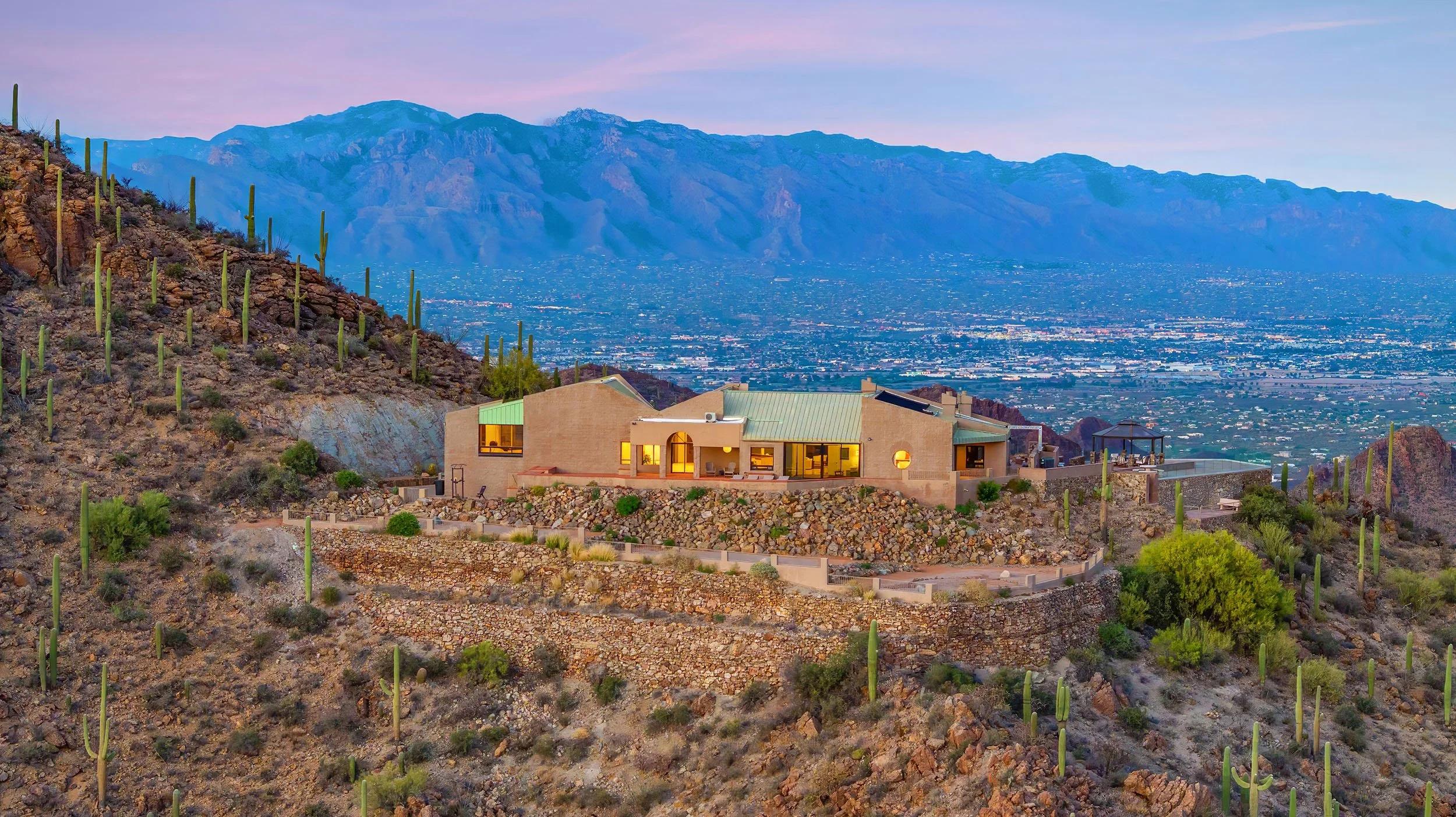 A modern house with large windows and a green roof situated on a hillside. The background features a vast cityscape with mountains and a colorful sky at sunset.