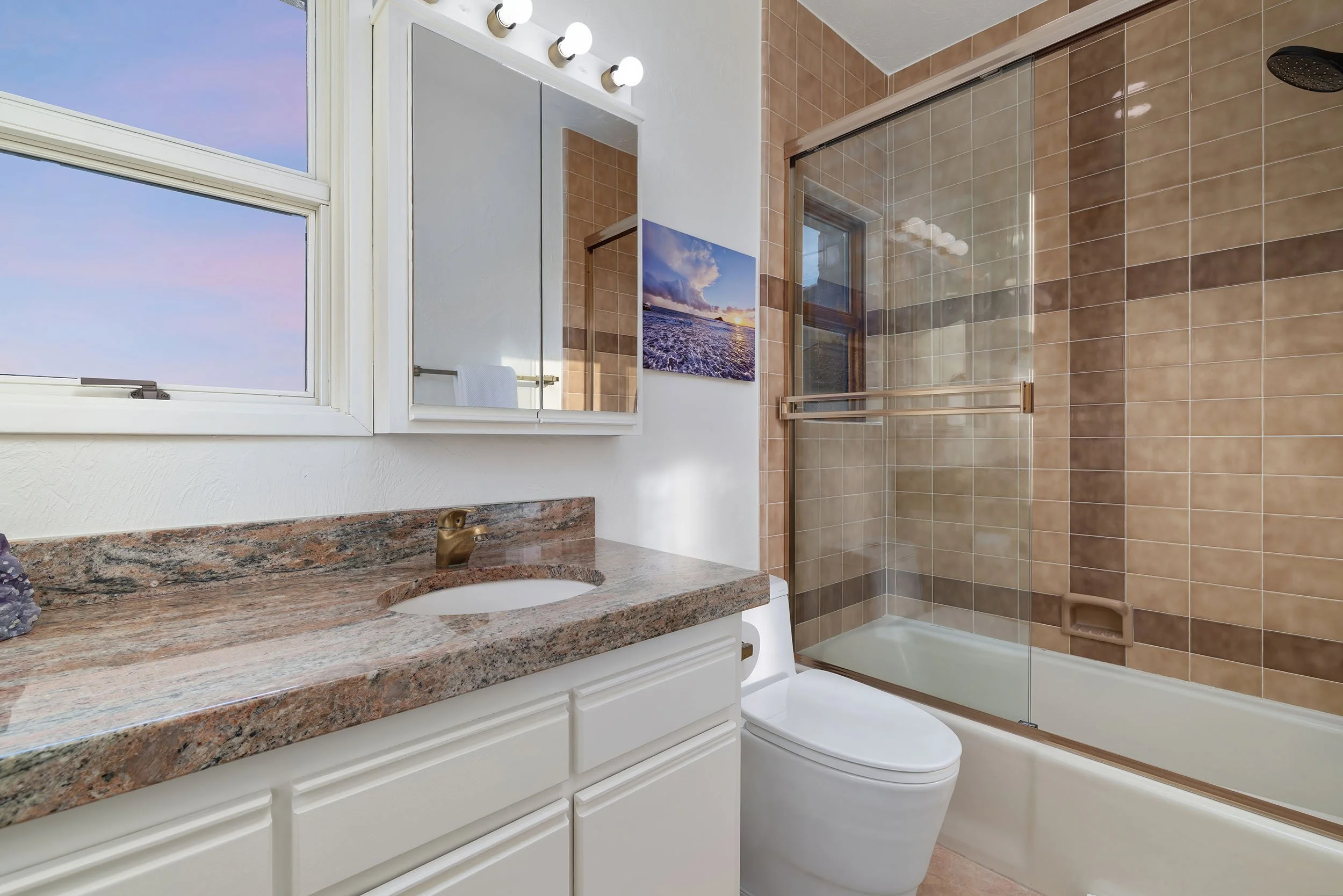 Bathroom with beige tiled shower, white vanity with granite countertop, mirror, window, and wall art of a beach scene.