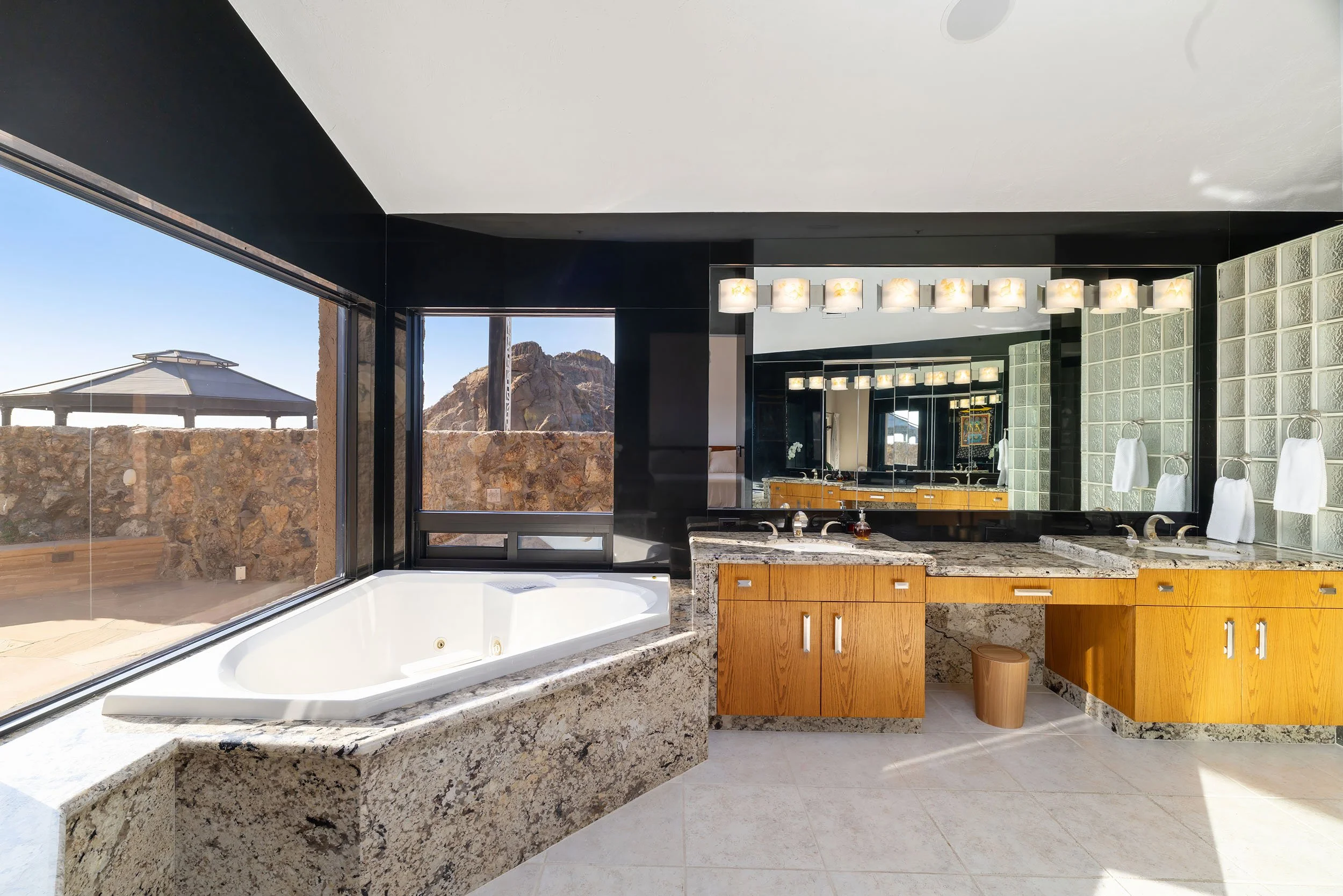 Modern bathroom with a large white bathtub, granite countertops, a wooden vanity, and a large mirror. There are glass block walls, towel hooks with white towels, and a window with a mountain view outside.