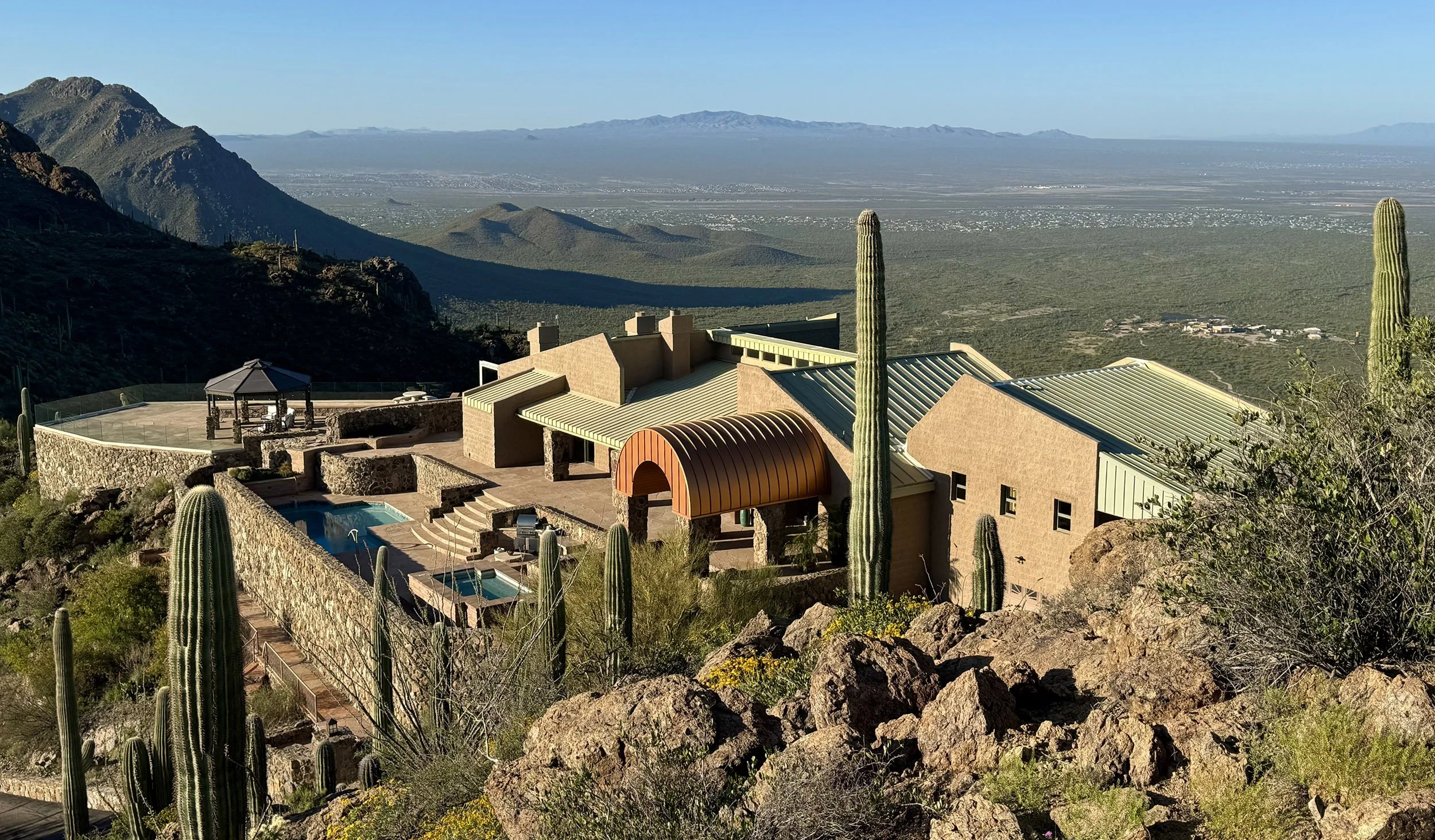 A house with desert landscaping, including tall cacti, situated on a mountainside overlooking a vast desert valley and mountain range in the distance.