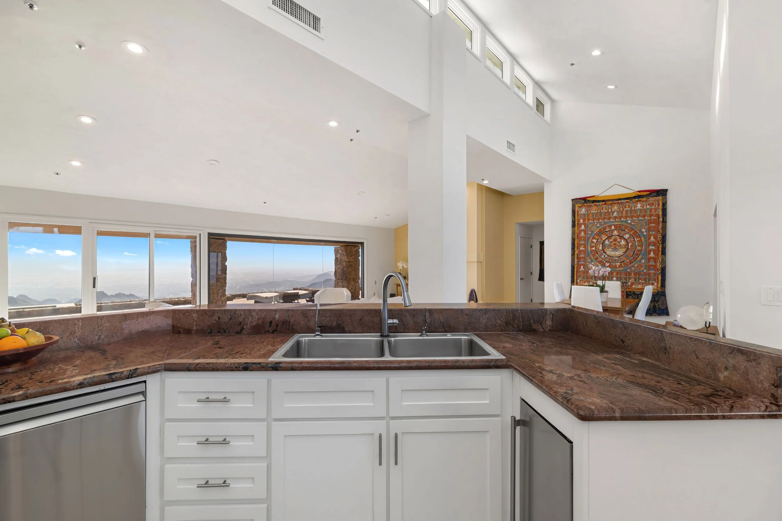 A modern kitchen with a granite countertop island featuring a double sink. Large windows overlook mountains. White cabinets and a colorful decorative wall hanging are visible.