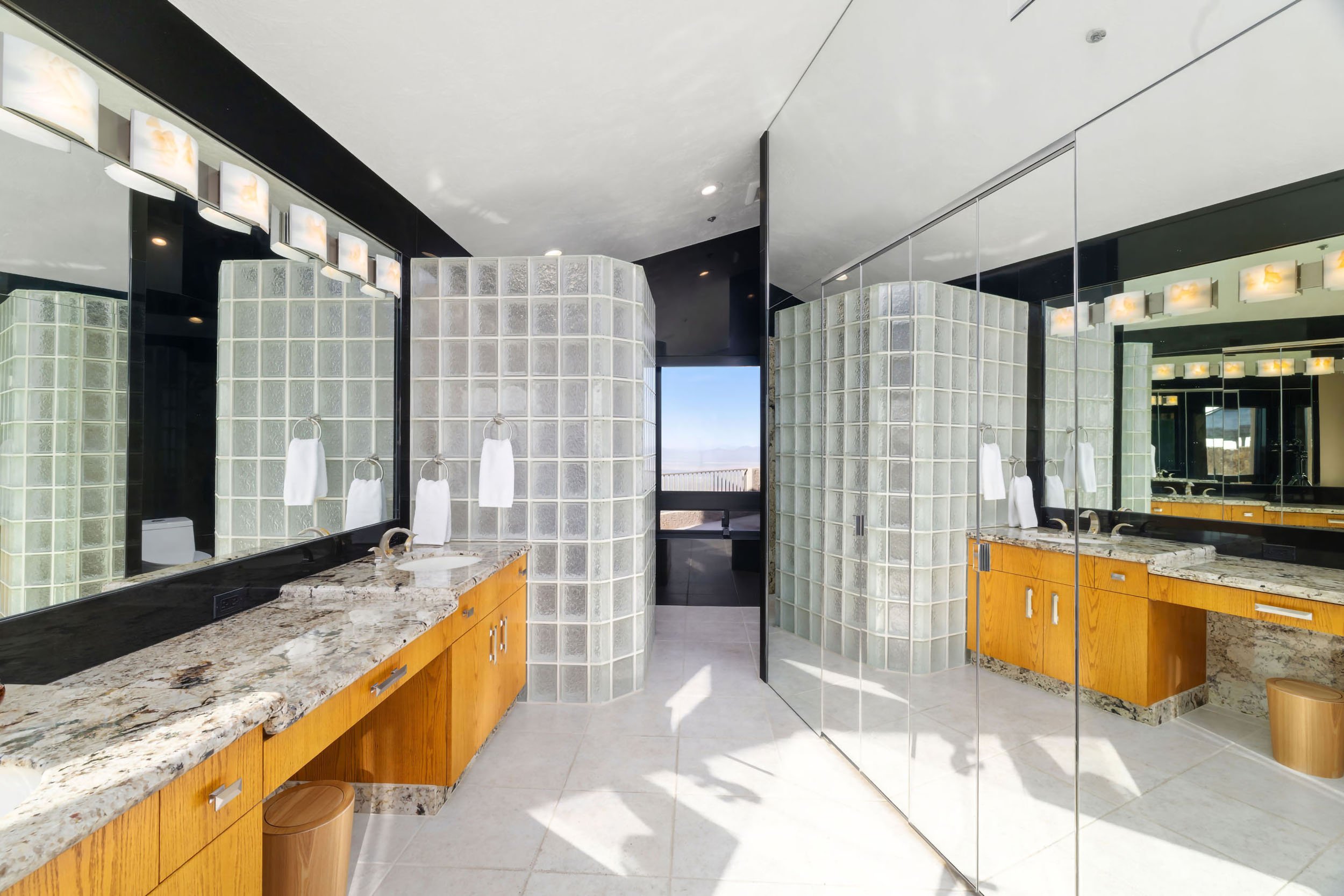 Modern bathroom with glass block walls, wooden cabinets, granite countertops, large mirrors, and multiple light fixtures.