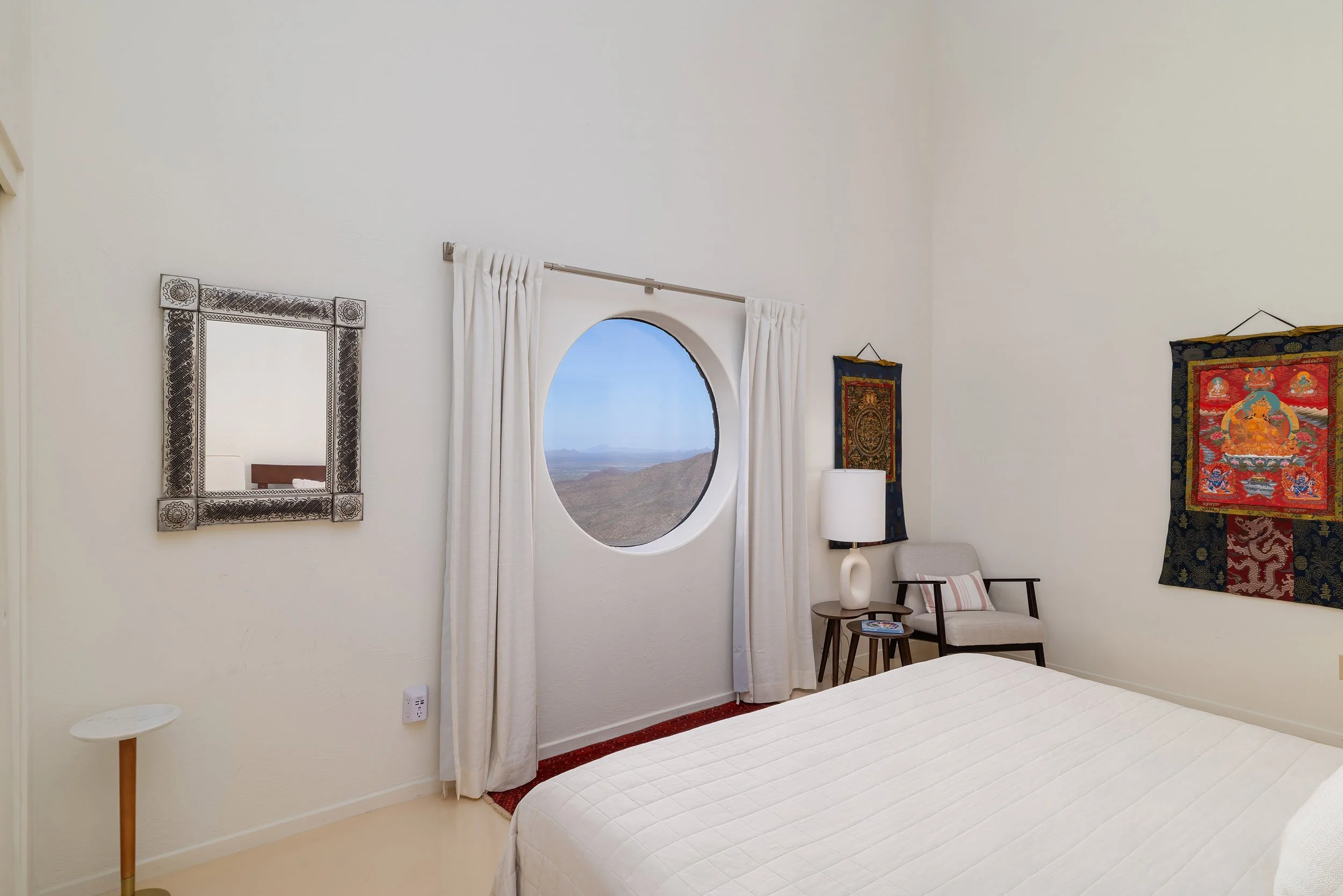 A bedroom with white walls, a round window showing a desert landscape, and colorful Asian tapestries on the wall. There is a bed with a white quilt, a beige armchair with a pillow, a white lamp, and a small side table.