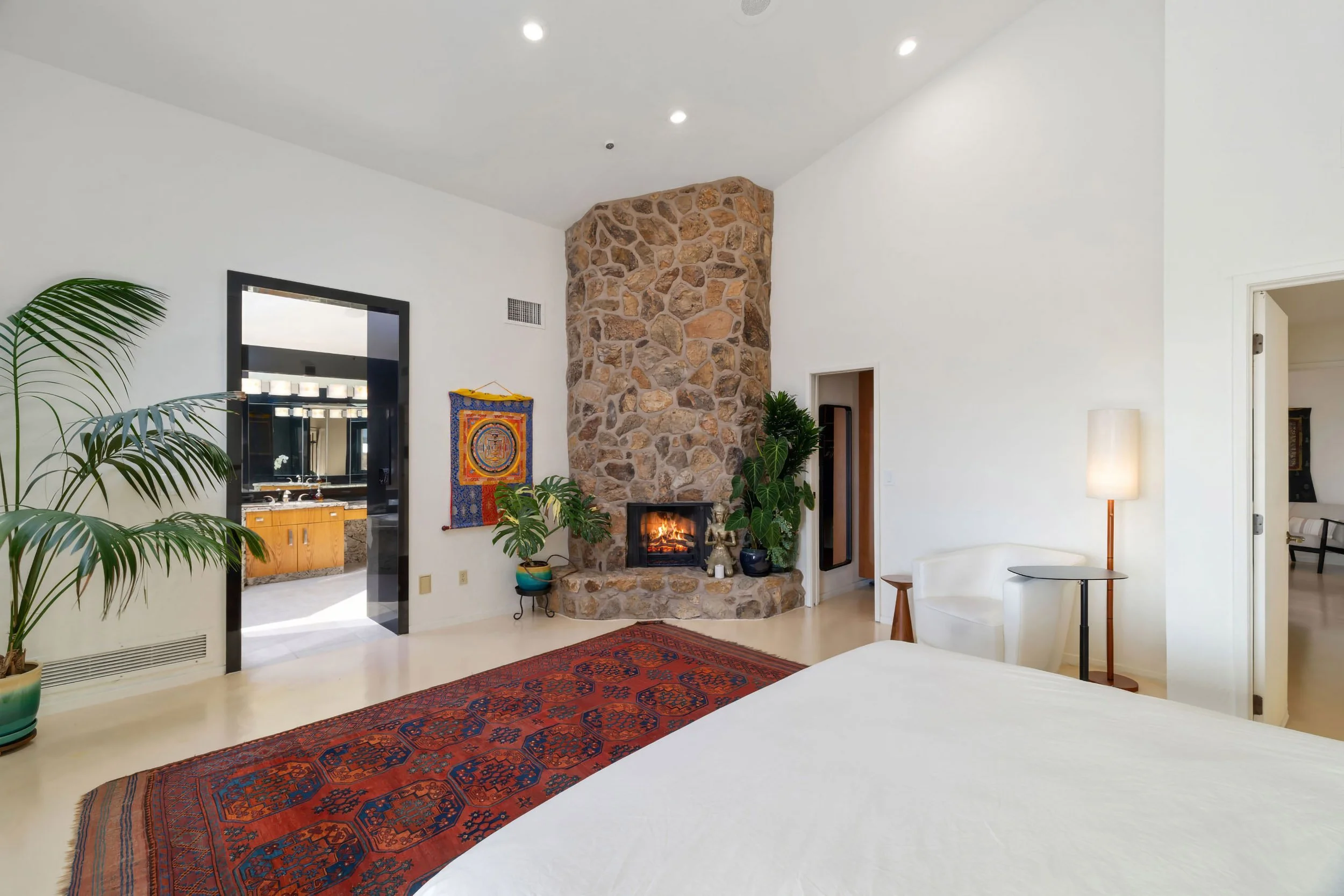 Living room with a stone fireplace, potted plants, a red rug, and a white chair next to a tall floor lamp, with an open doorway leading to a bedroom.