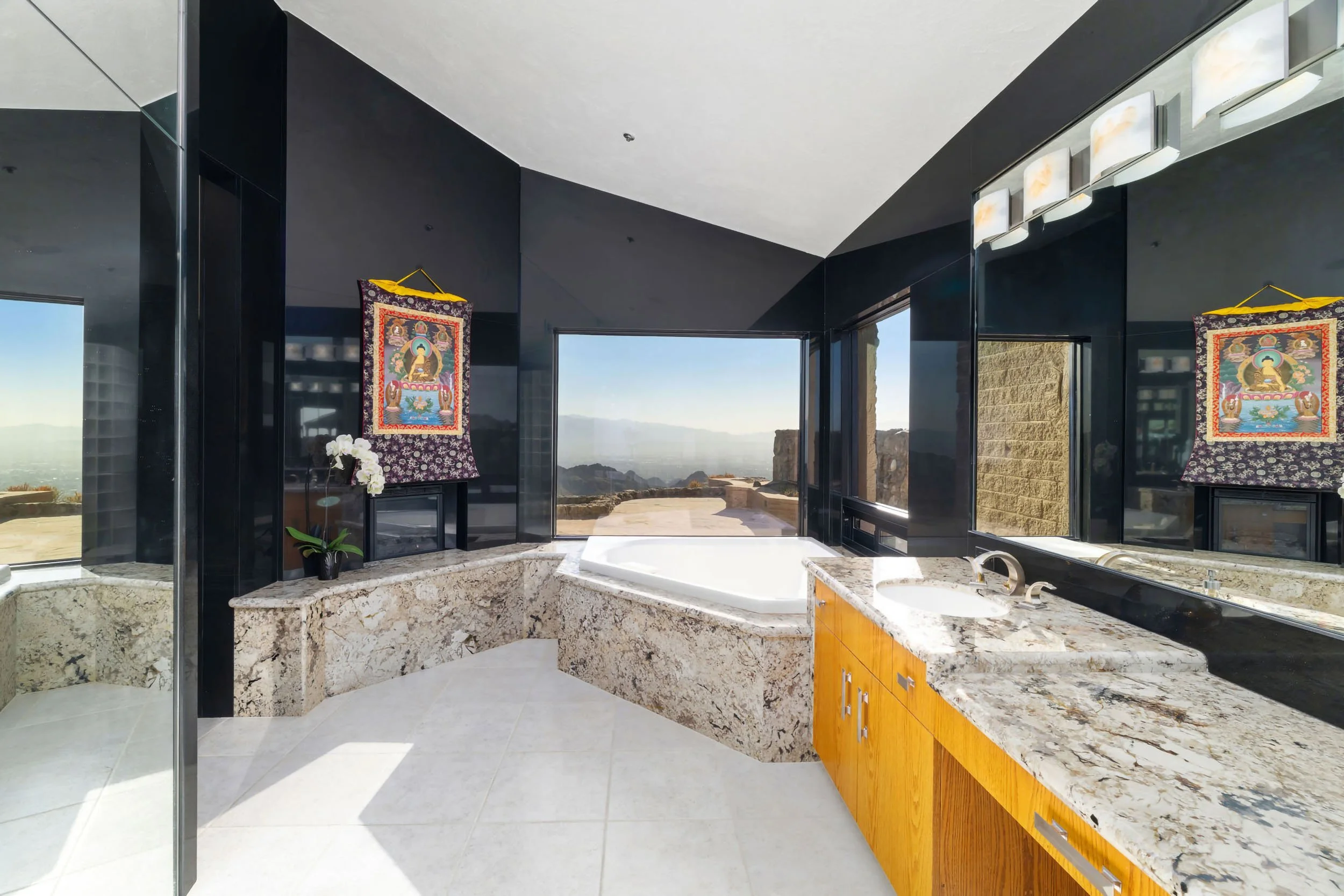 Modern bathroom with granite countertops, a bathtub, and large windows showing an outdoor view, with religious artwork hanging on the dark wall.