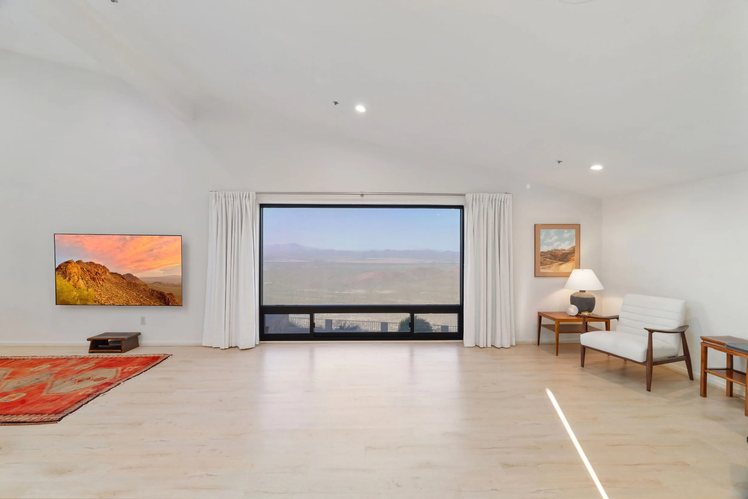 Living room with large window showing a desert landscape, a wall-mounted TV displaying a sunset over mountains, a white armchair, wooden side tables, a lamp, framed landscape photo, and a rug on light-colored wooden floor.