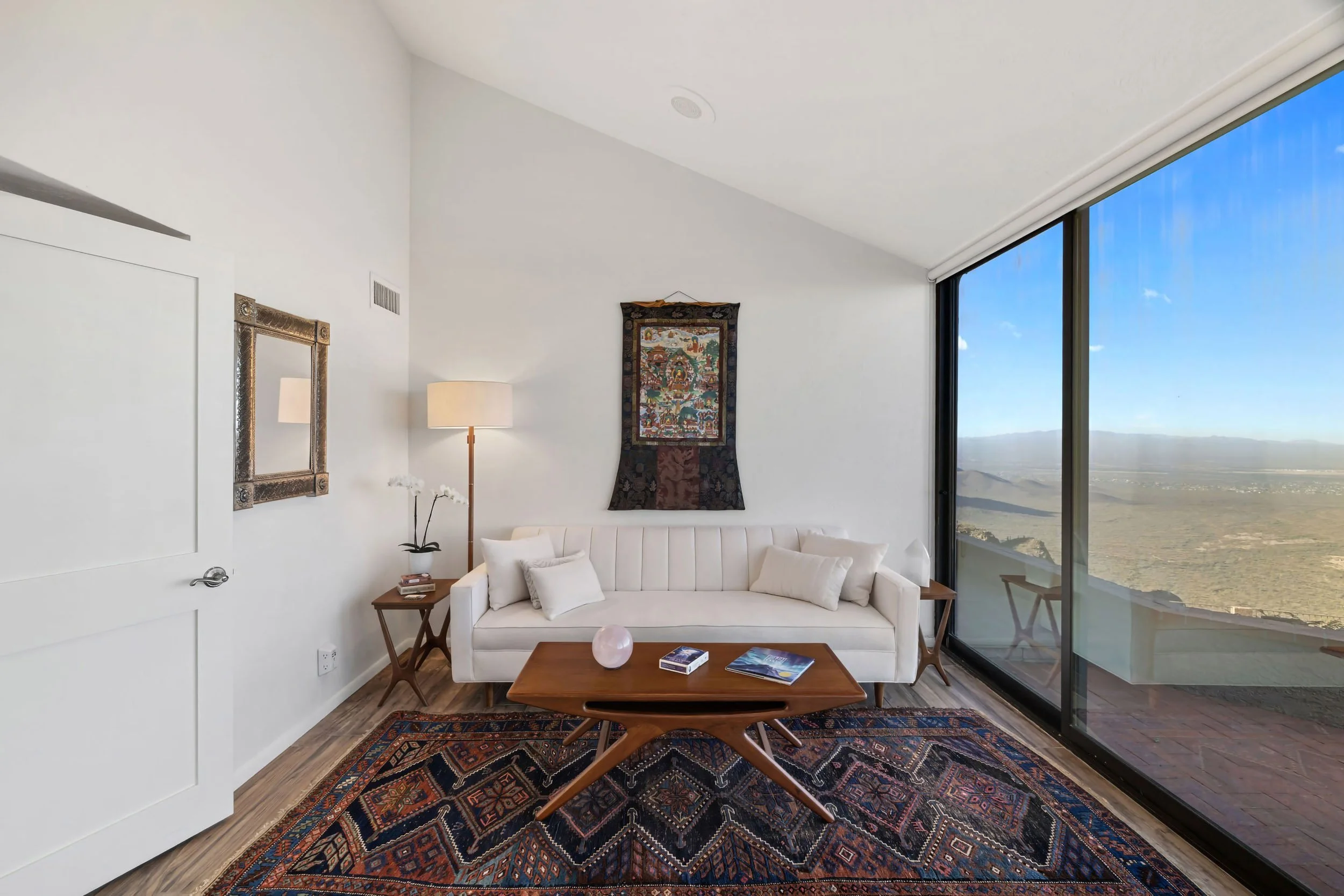 A living room with white walls, a white sofa with pillows, a wooden coffee table with magazines, a patterned rug, a floor lamp, wall art, and large windows showing an outdoor landscape.