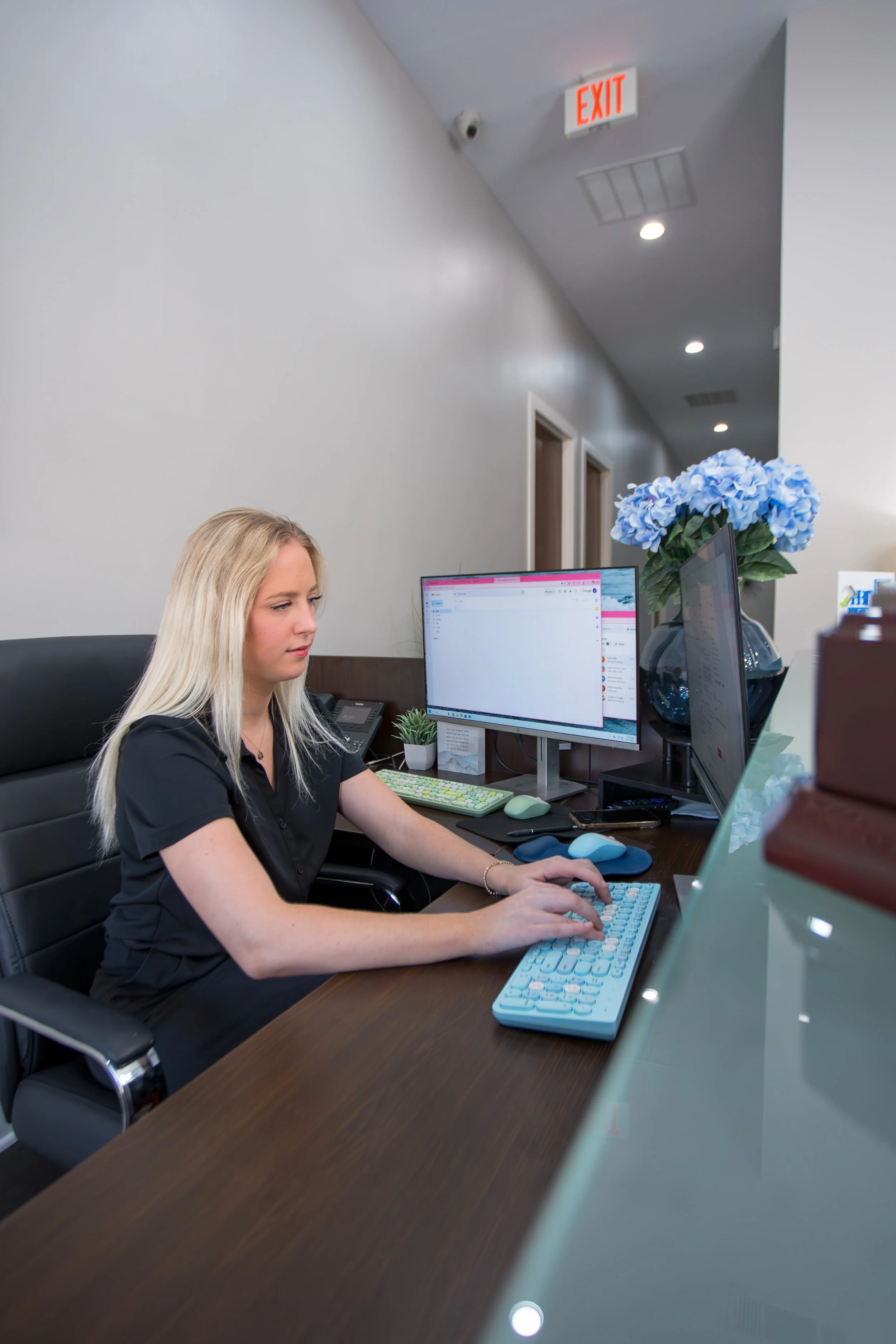A woman with blonde hair working at a desk in an office, using a large computer keyboard with a light blue cover, with dual monitors, a plant, and a large vase of blue hydrangeas on her right.