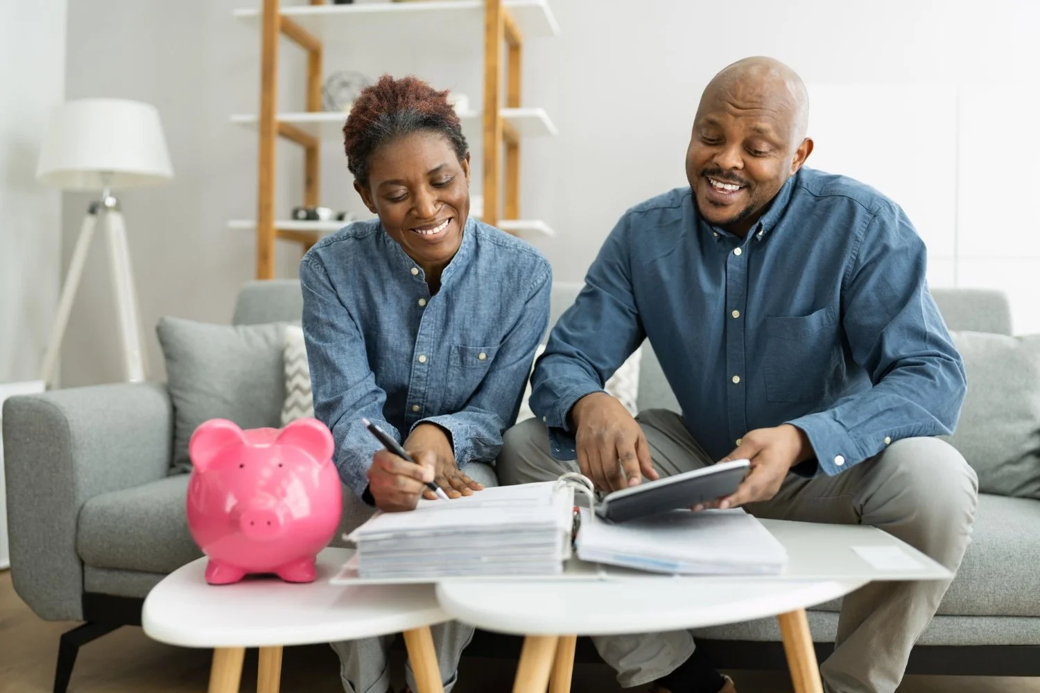 A happy couple sitting on a gray sofa at home, reviewing financial documents on a white coffee table with a pink piggy bank nearby.