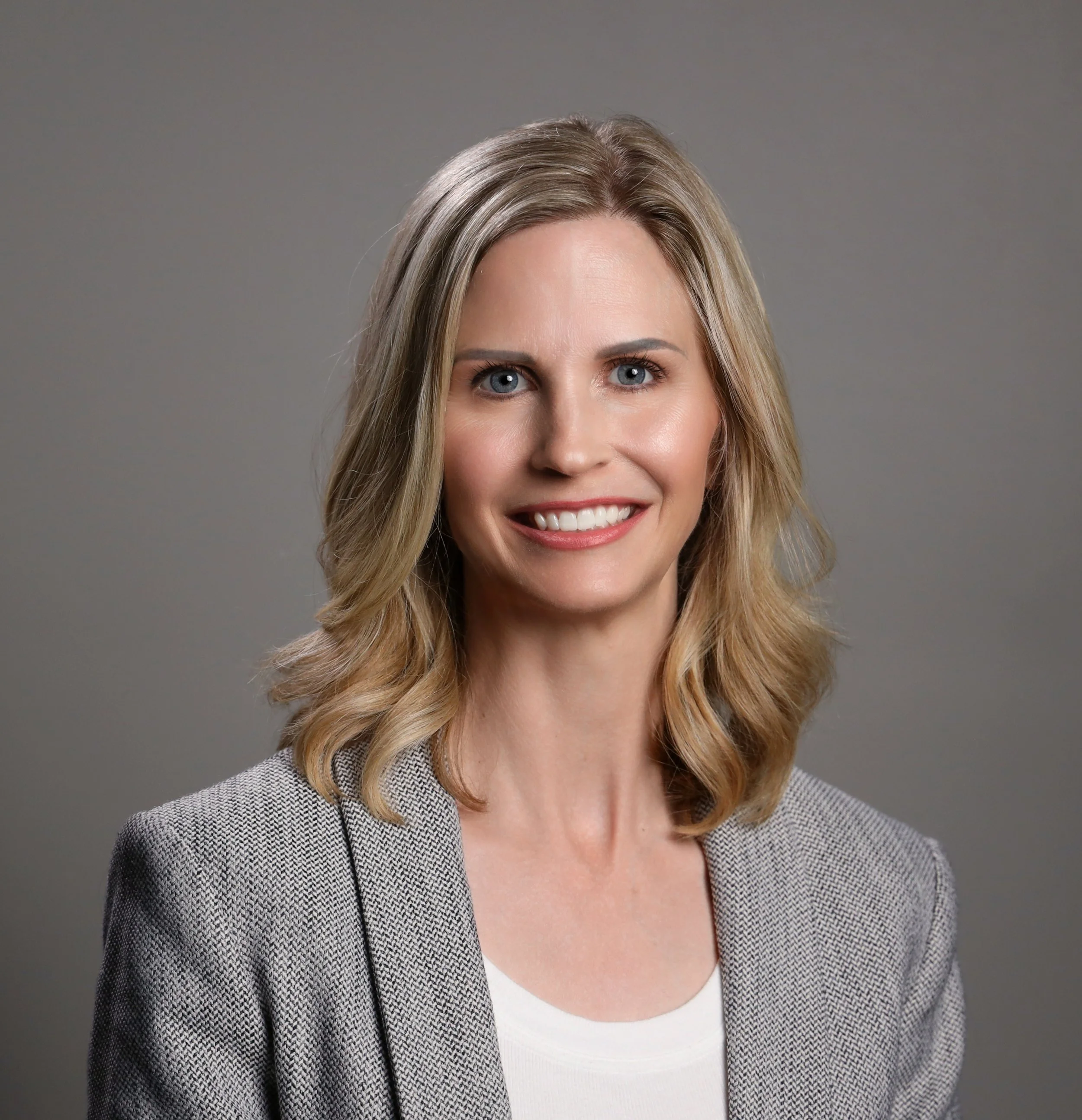 Close-up portrait of a smiling woman with blonde hair wearing a gray checked blazer over a white top against a gray background.