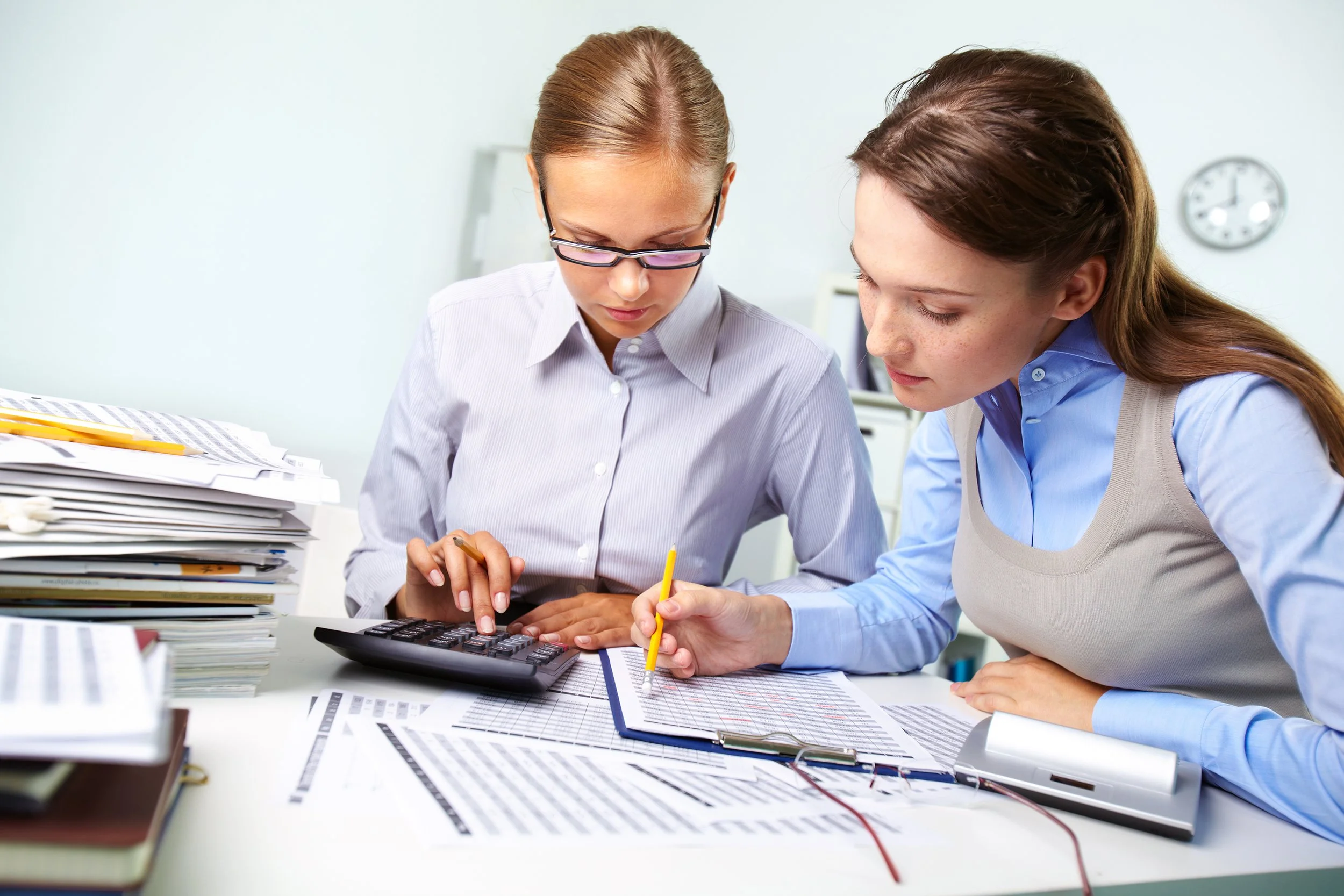 Two women working together at a desk with documents, a calculator, and writing tools, in an office setting.