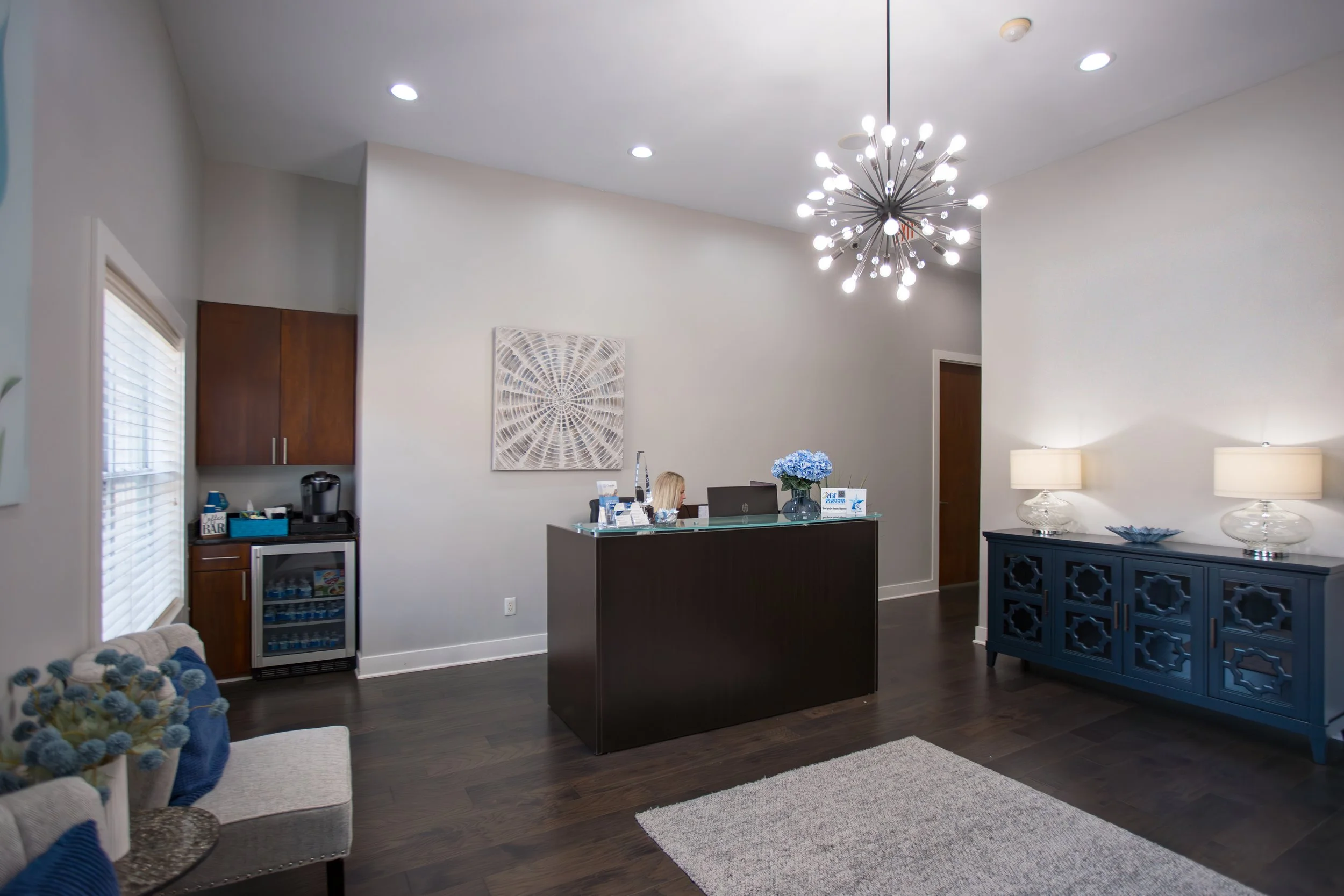 A modern reception area with white walls, dark hardwood floors, a black reception desk with a woman working, a round chandelier with multiple small lights, a blue cabinet with decorative items, a small refrigerator, and a seating area with a sofa and a basket of blankets.