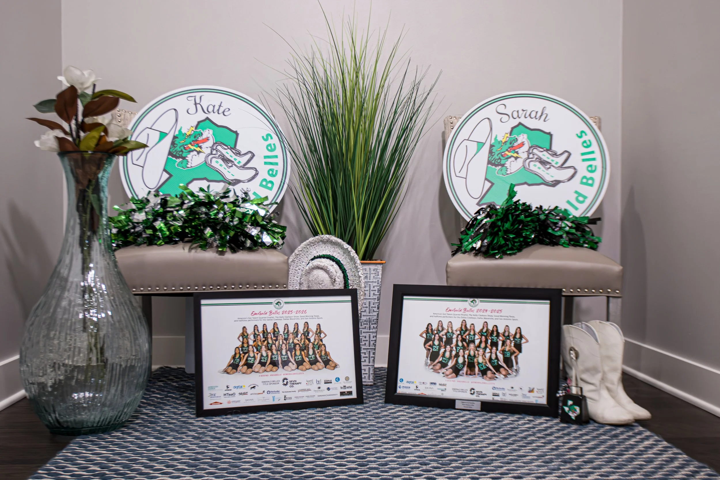 Decorative display with two chairs, each with a sign for 'Esmarla Bellies' and the names Kate and Sarah, featuring a cartoon mascot of a dragon and sneakers. A large green plant is centered between the chairs. The display includes a glass vase with artificial flowers, a small rainbow decoration, framed photos of cheerleaders, and various sponsor logos on the photos.