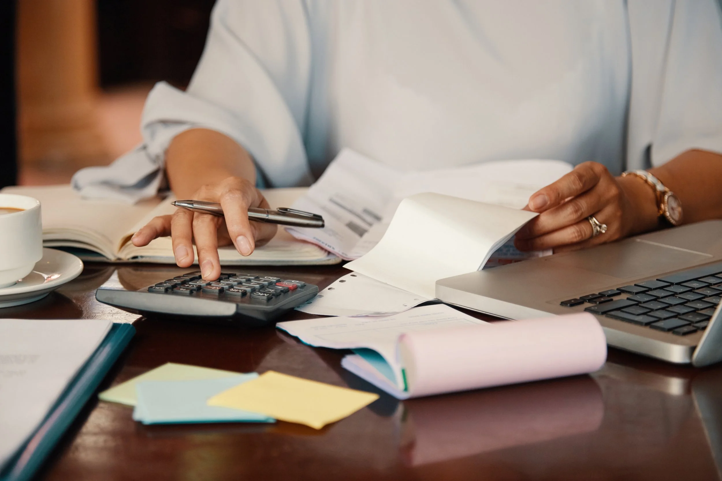 A person in a white shirt working at a cluttered desk with a calculator, open book, papers, folders, a pen, a laptop, and a cup of coffee.