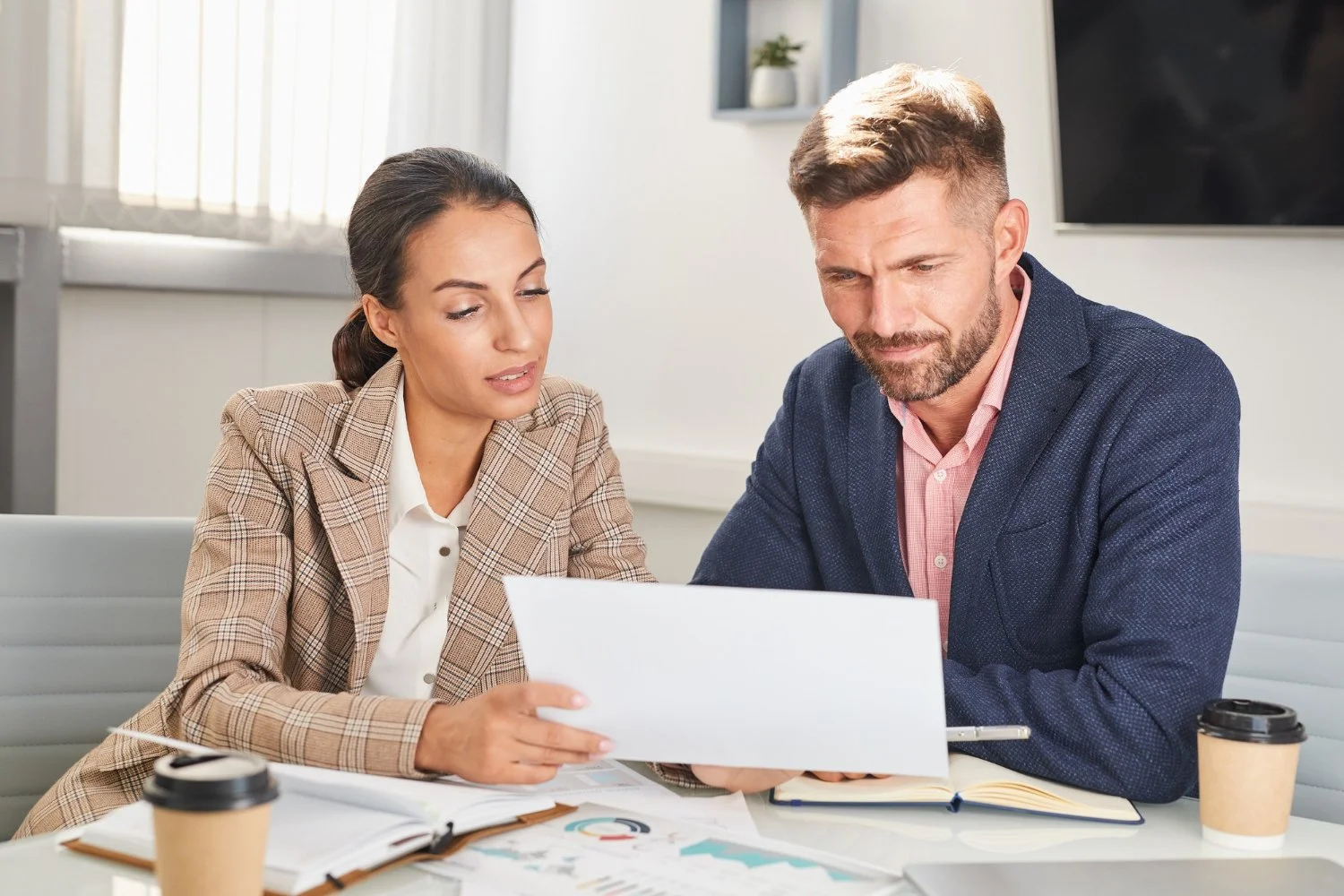 A man and a woman in business attire reviewing documents at a table in an office with coffee cups.