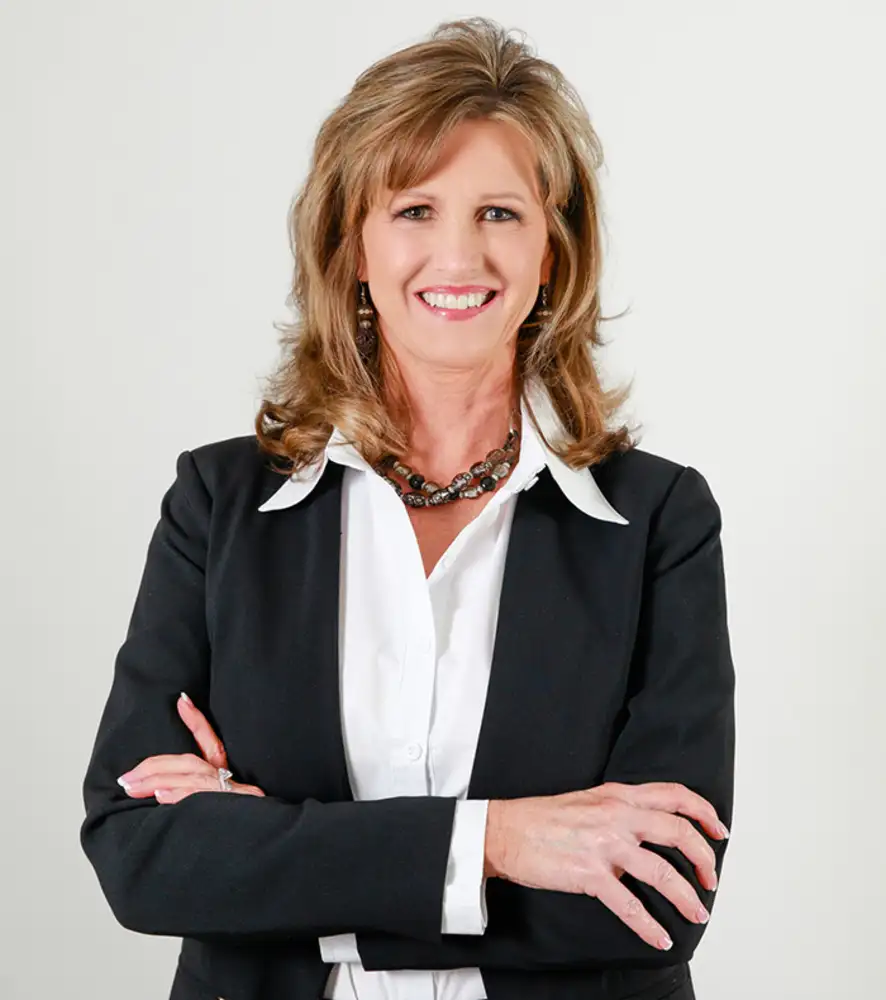 A woman with shoulder-length blonde hair, wearing a black blazer over a white shirt, accessorized with a necklace and earrings, standing with her arms crossed and smiling against a plain light background.