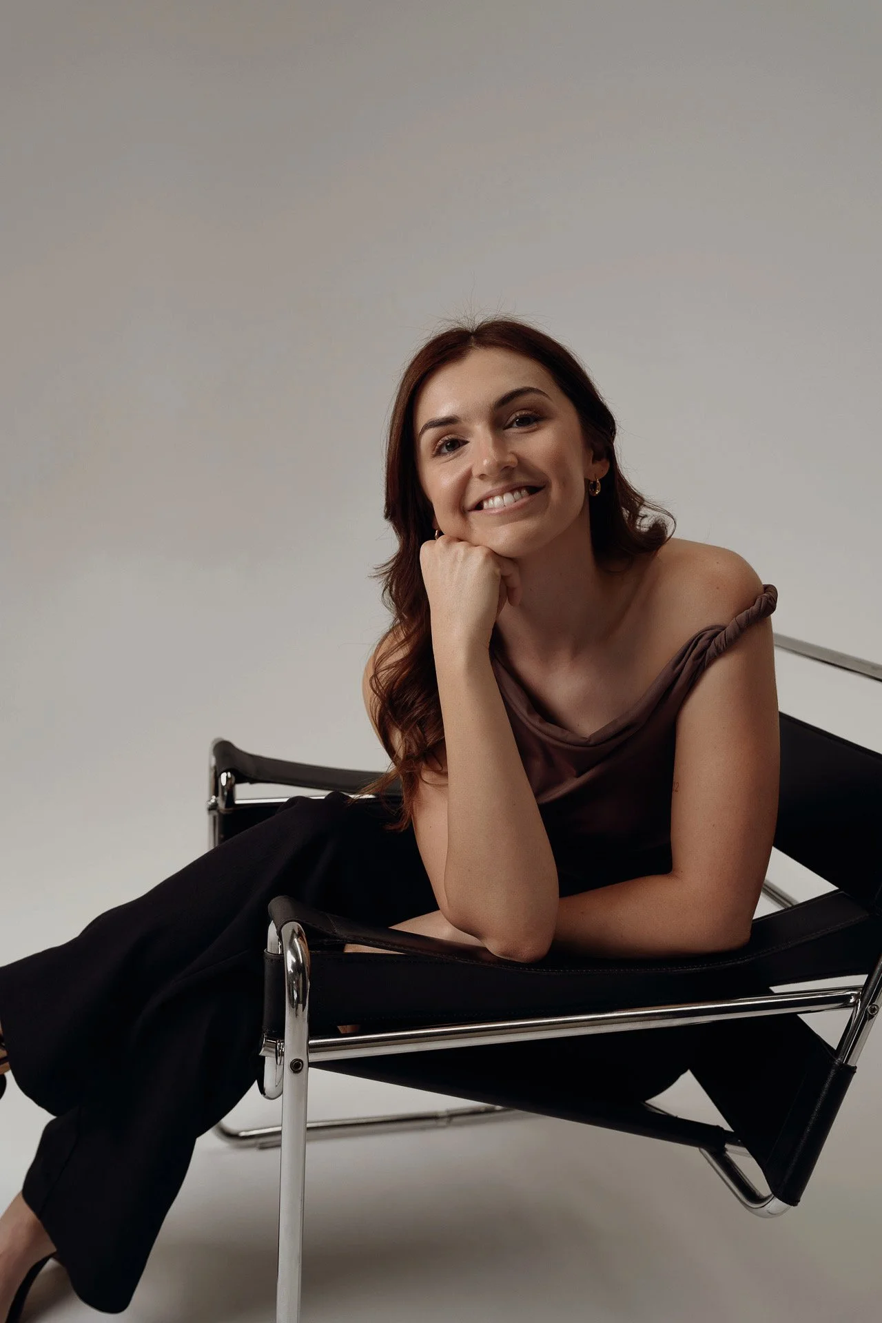 A woman with brown hair, wearing a sleeveless brown top and black pants, reclining on a black lounge chair with chrome armrests, smiling at the camera against a plain light gray background.