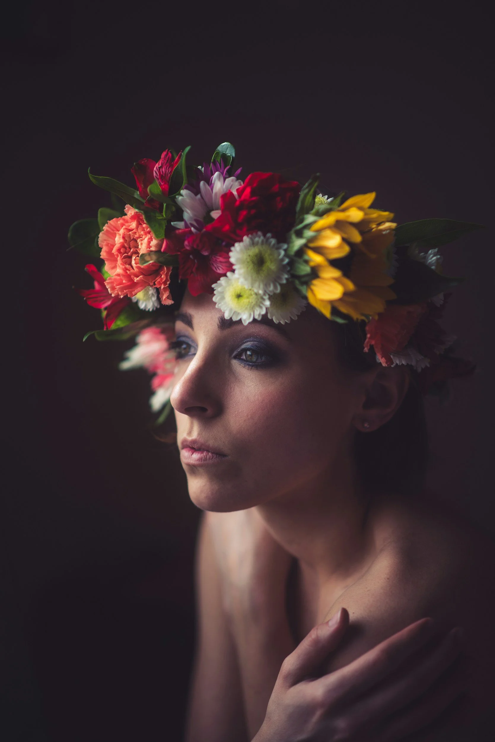 A woman with a floral crown made of colorful flowers and green leaves, looking to the side with a thoughtful expression against a dark background.